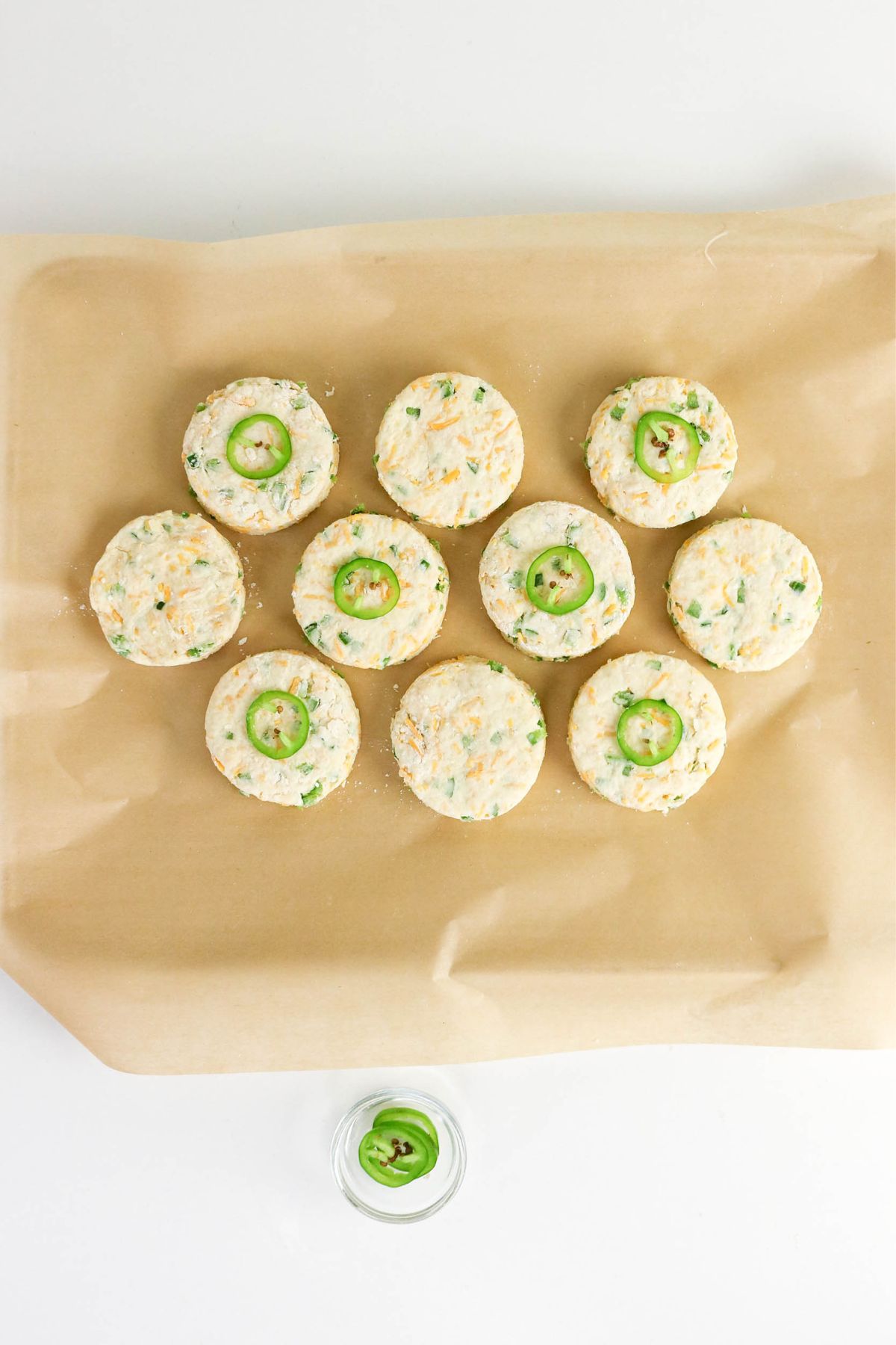 Ten round, unbaked biscuits with green jalapeño slices on top are arranged on parchment paper; a small glass bowl of green sauce sits nearby.