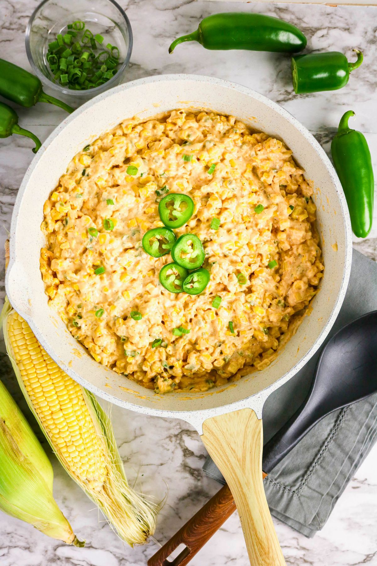 A skillet of creamy corn with green jalapeño slices on top, surrounded by fresh corn, whole jalapeños, and a bowl of chopped green onions on a marble surface.