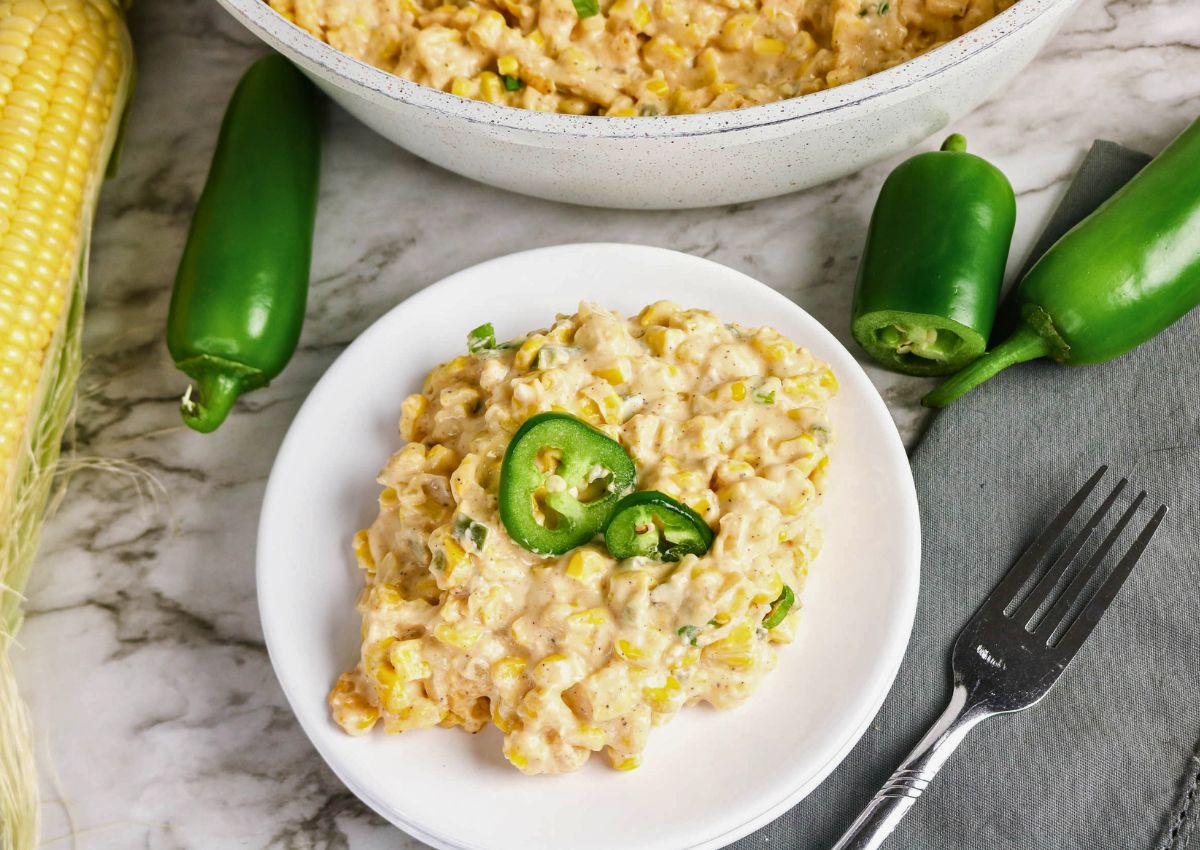 A plate of creamy corn casserole topped with jalapeño slices, next to fresh jalapeños, a fork, and corn on the cob on a marble surface.