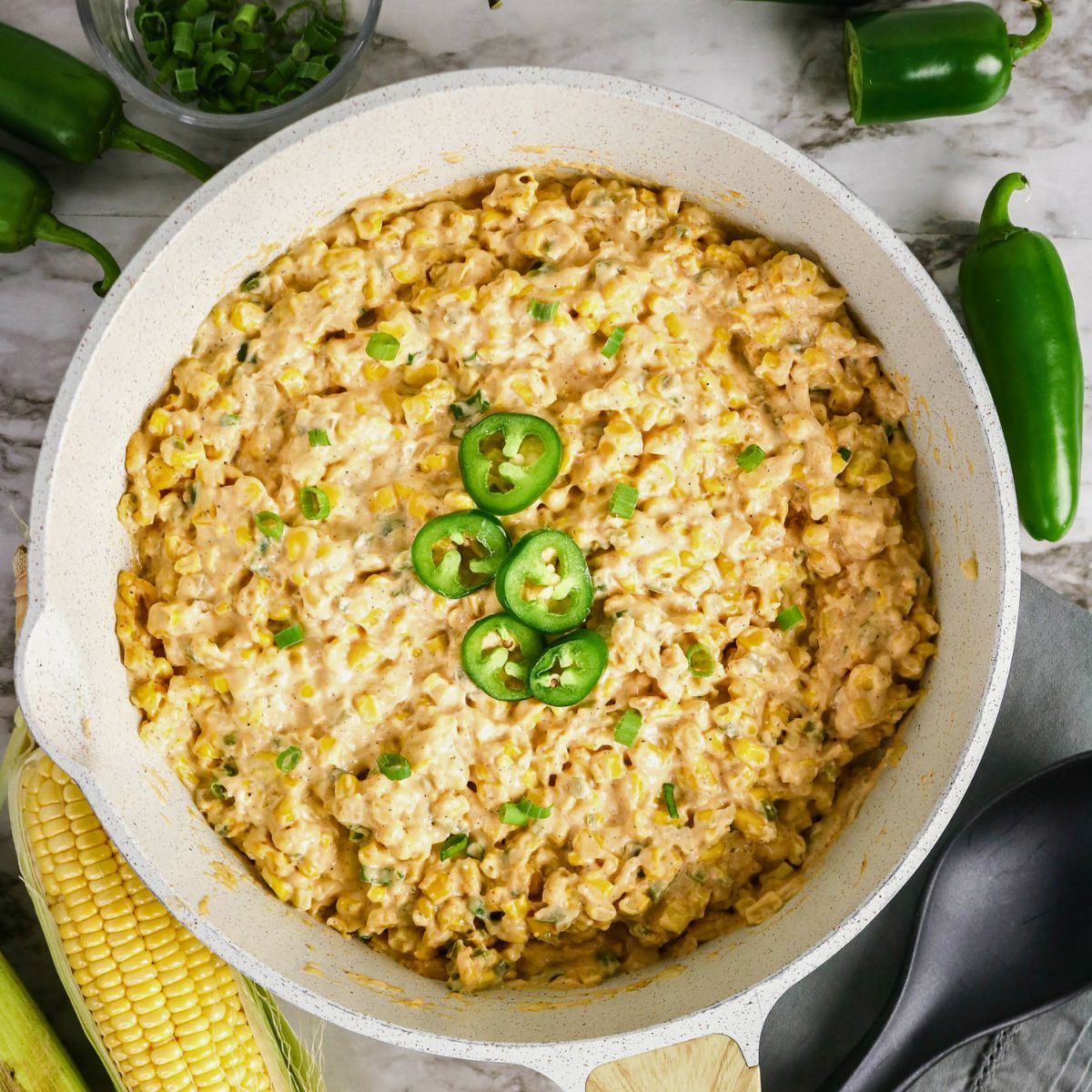 A skillet filled with creamy corn dip topped with sliced jalapeños and chopped green onions, surrounded by fresh corn and whole jalapeños on a marble surface.