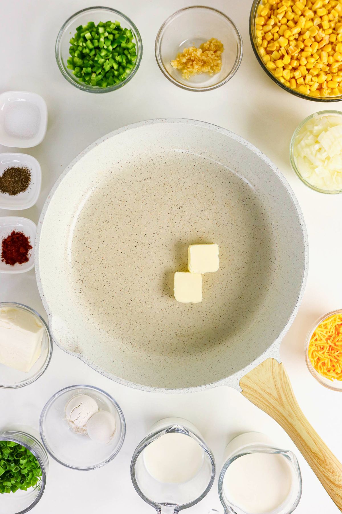 A skillet with two butter cubes is surrounded by bowls of chopped vegetables, seasonings, corn, cheese, cream, flour, and milk on a white surface.