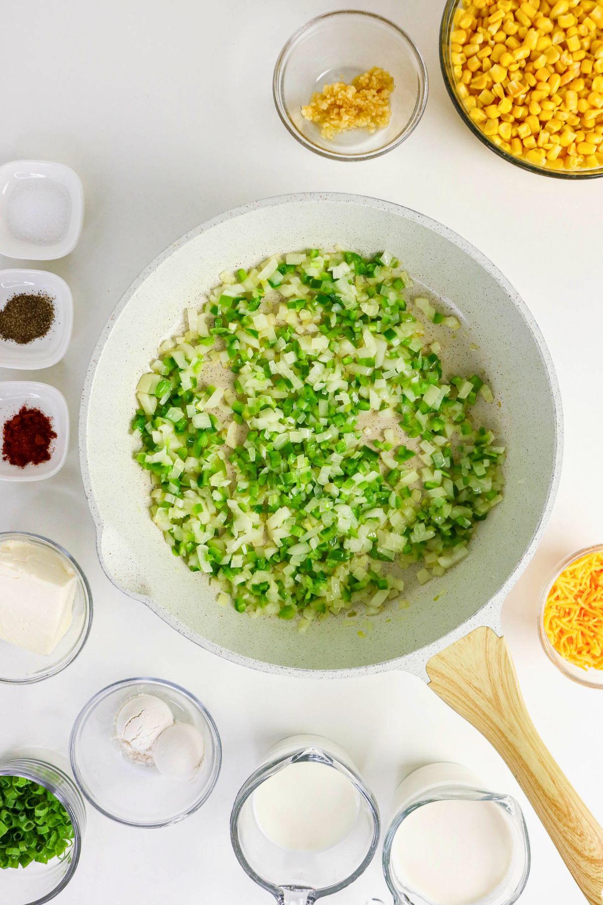 A skillet with chopped green bell peppers, celery, and onions, surrounded by small bowls of corn, cheese, seasonings, milk, flour, and butter on a white surface.
