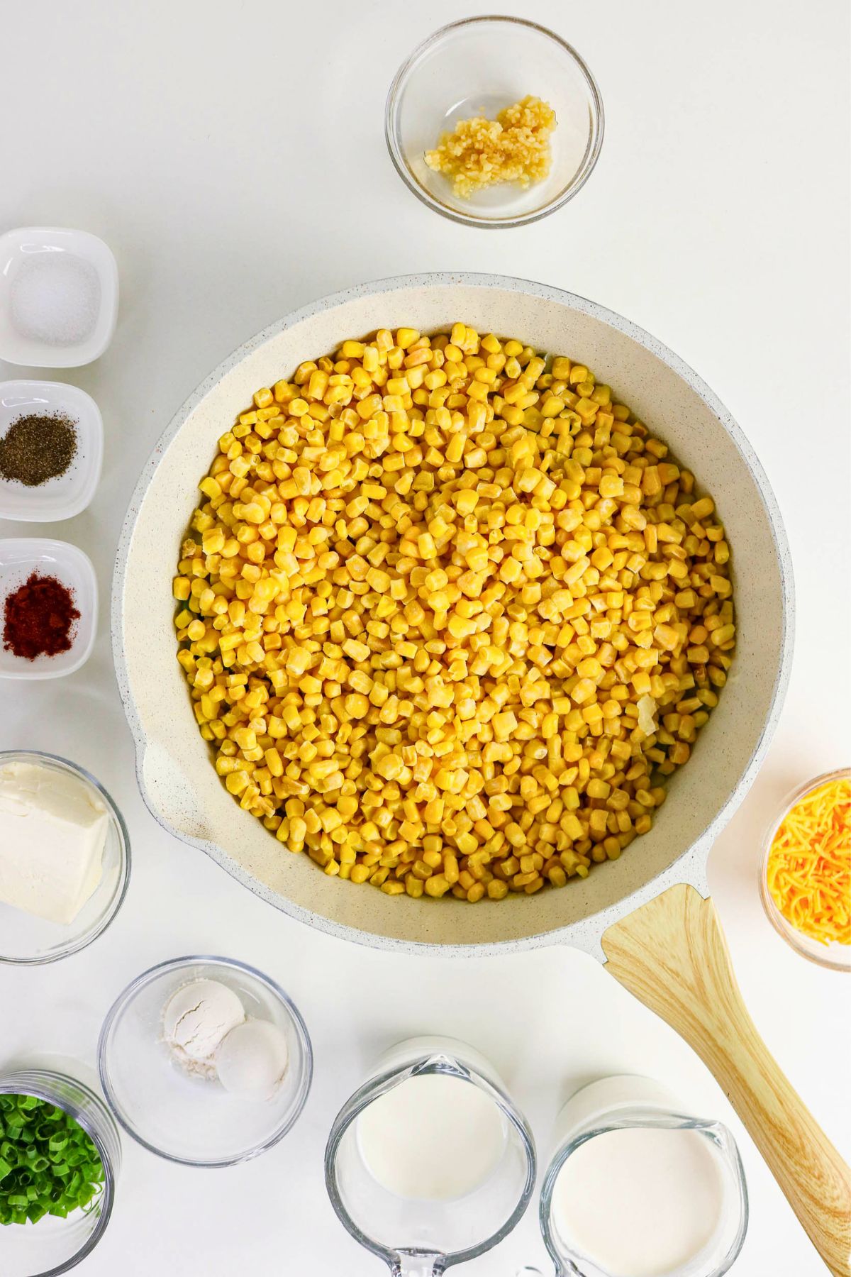 A white pan filled with corn kernels is surrounded by small bowls containing spices, garlic, cream, milk, flour, green onions, and shredded cheese on a white surface.