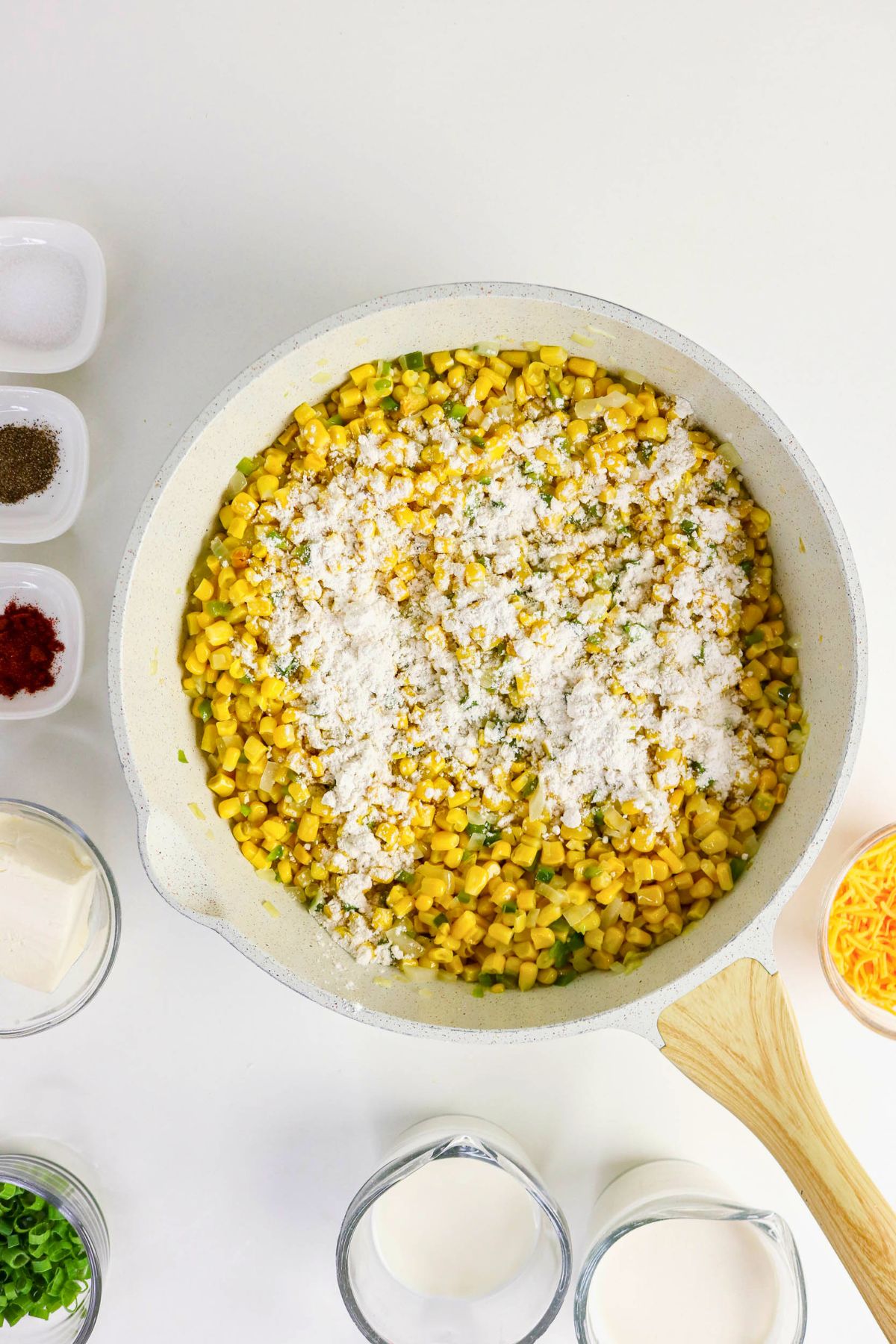 A skillet with corn, spices, and chopped herbs topped with flour, surrounded by small bowls of shredded cheese, spices, cream, and green onions on a white surface.