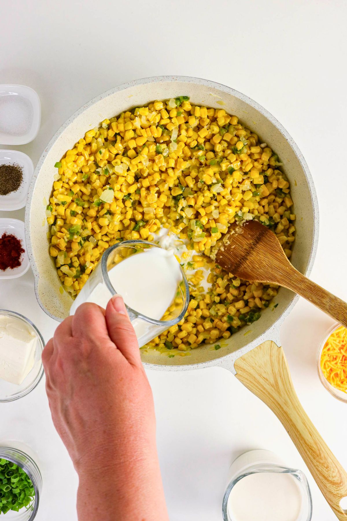 A hand pours milk into a pan of sautéed corn and green peppers, surrounded by bowls of ingredients on a white surface.