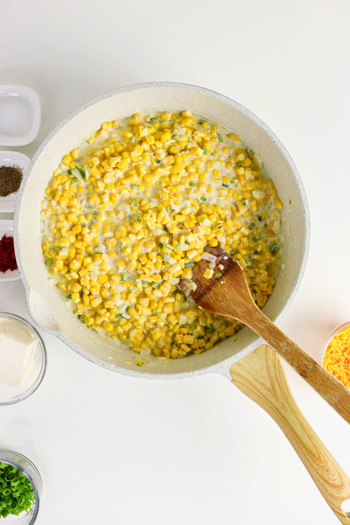 Corn and chopped green onions being stirred in a creamy sauce in a white skillet, with a wooden spoon and various ingredients in bowls nearby.