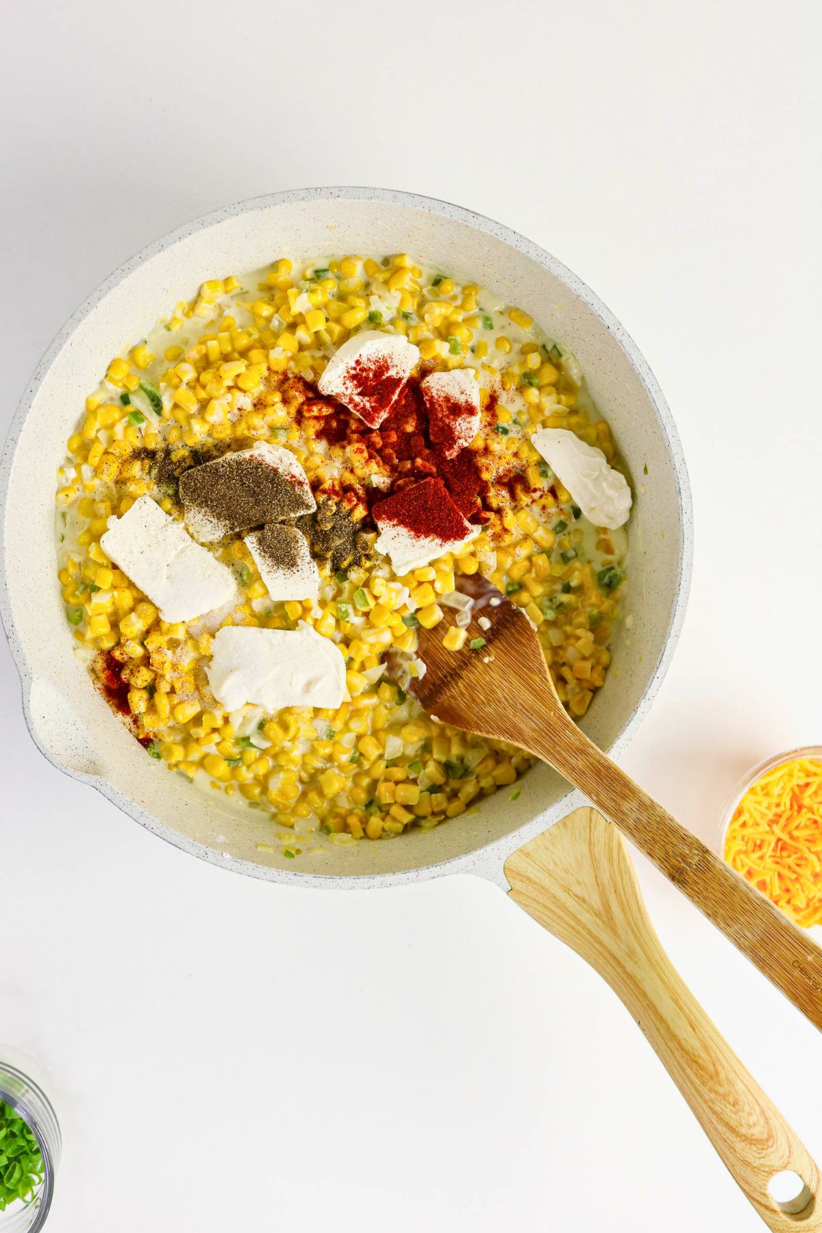 A skillet with corn, cream cheese, seasonings, and a wooden spoon, ready to be mixed, on a white surface.