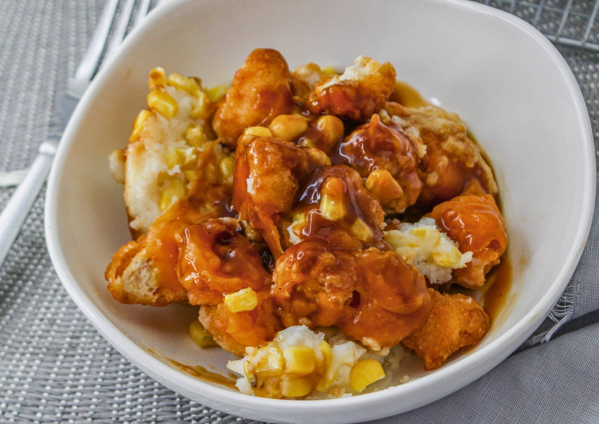 A white bowl filled with mashed potatoes, corn, popcorn chicken, and brown gravy sits on a gray placemat with a fork nearby.
