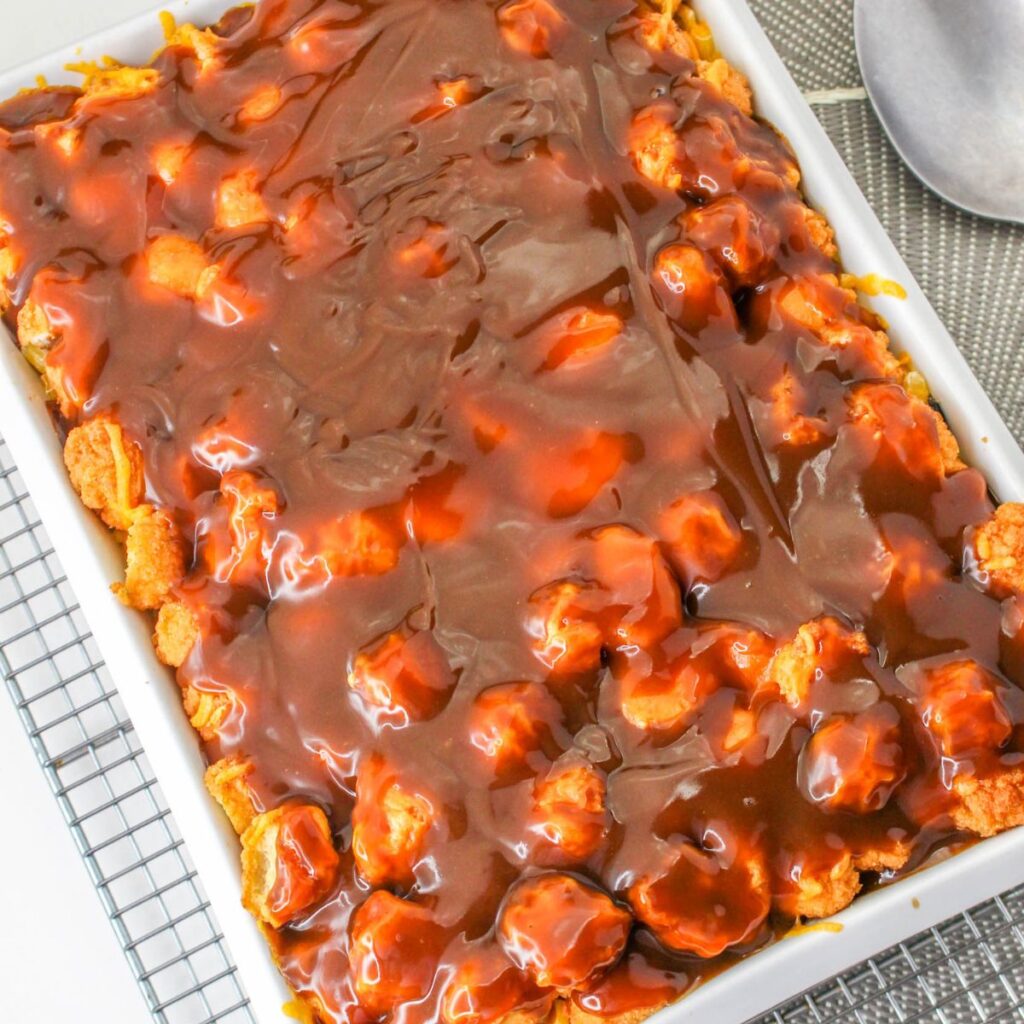 A rectangular baking dish filled with bread pudding topped with a glossy layer of chocolate sauce, placed on a cooling rack next to a metal serving spoon.