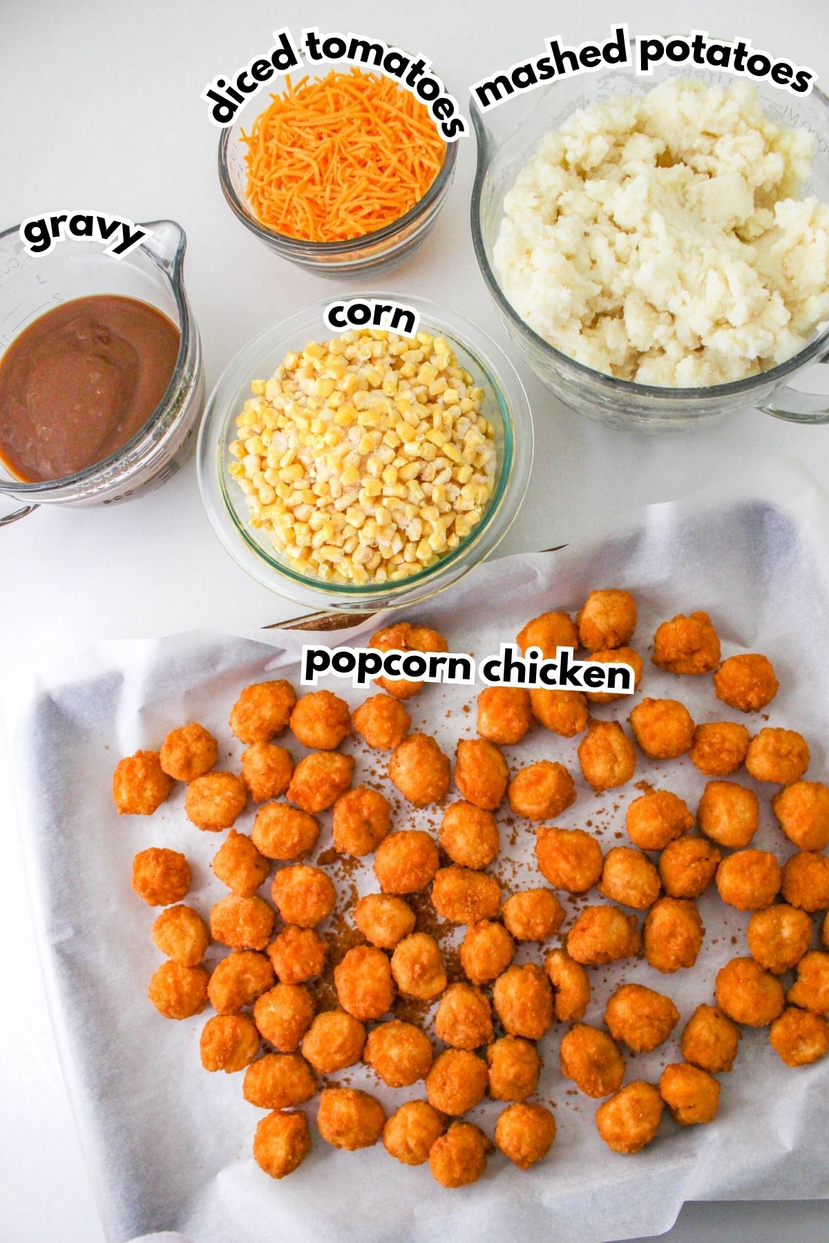 Bowls of diced tomatoes, mashed potatoes, corn, and gravy surround a tray of popcorn chicken arranged on parchment paper. Labels identify each ingredient.