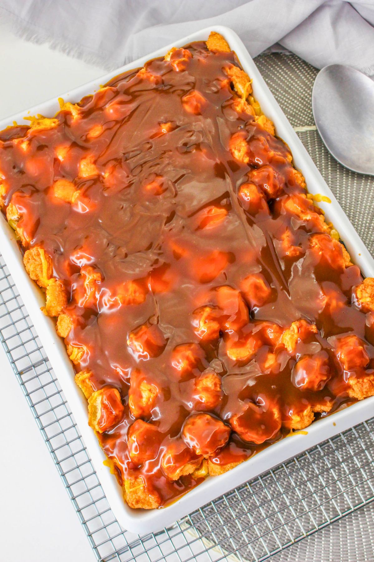 Rectangular baking dish filled with bread pudding topped with a glossy layer of chocolate sauce, placed on a wire cooling rack with a spoon nearby.