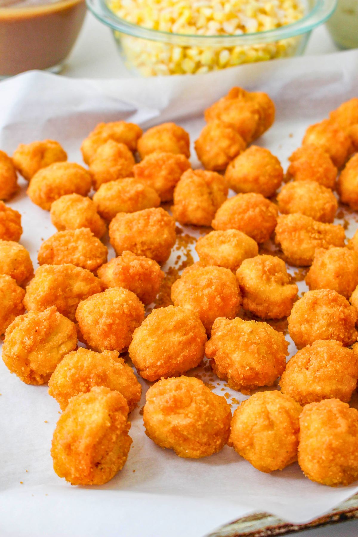 A tray lined with parchment paper holds several pieces of golden, breaded and fried chicken nuggets, with bowls of corn and sauce in the background.