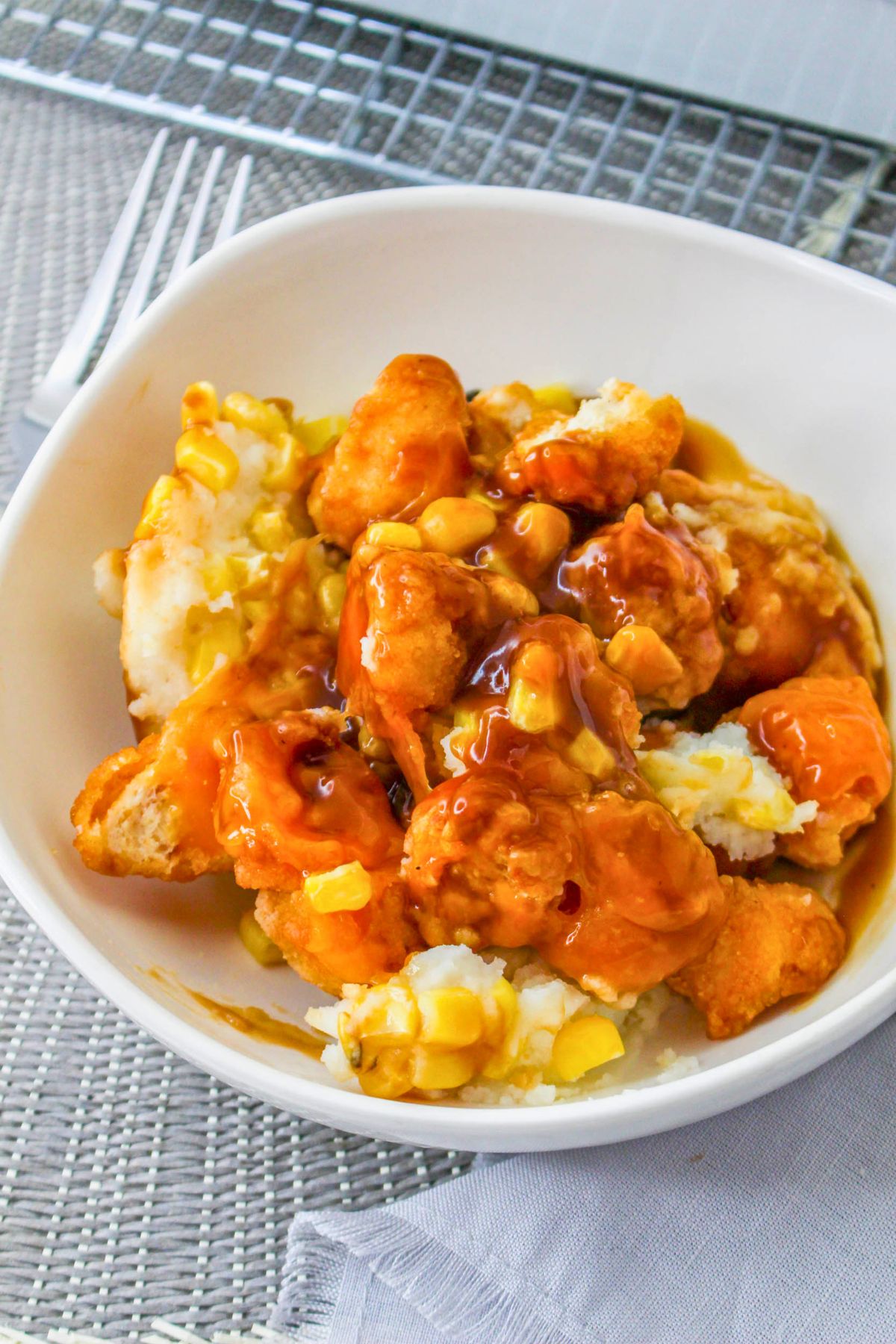 A white bowl filled with mashed potatoes, corn, breaded chicken pieces, and brown gravy on a woven placemat.