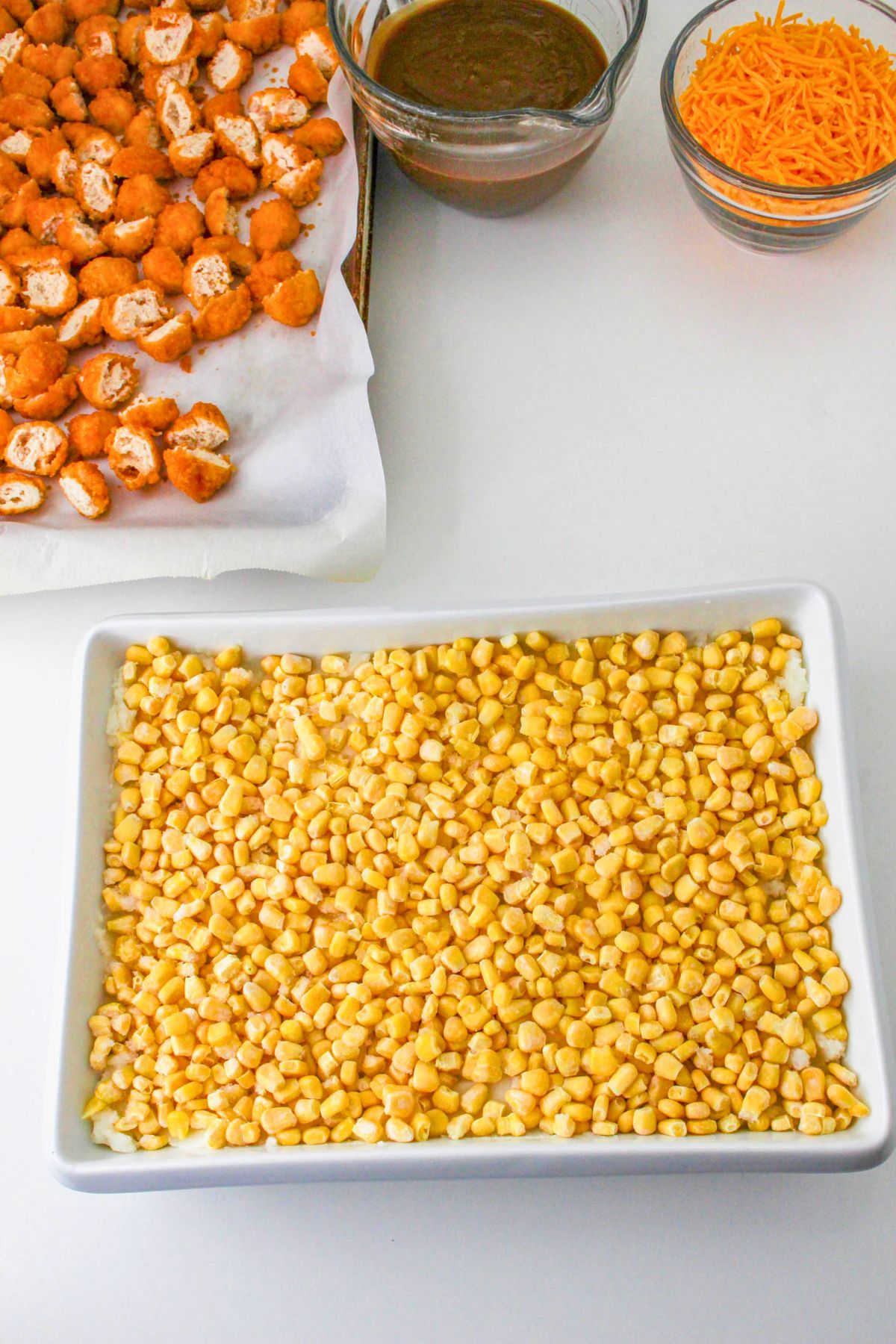 A baking dish filled with corn, a tray of breaded chicken pieces, a bowl of brown sauce, and a bowl of shredded cheese on a white surface.
