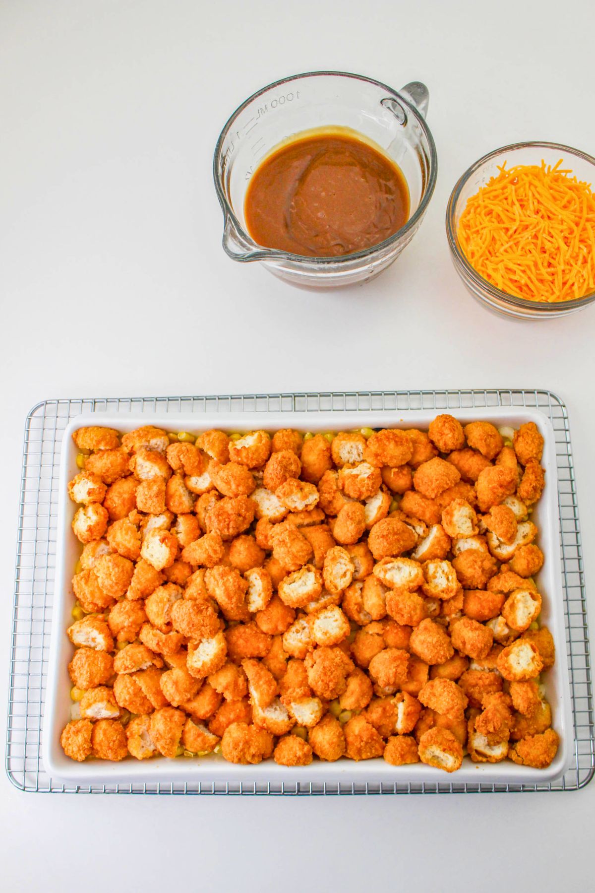Tray of breaded chicken pieces on a wire rack, with a bowl of shredded cheddar cheese and a glass measuring cup containing brown sauce nearby on a white surface.