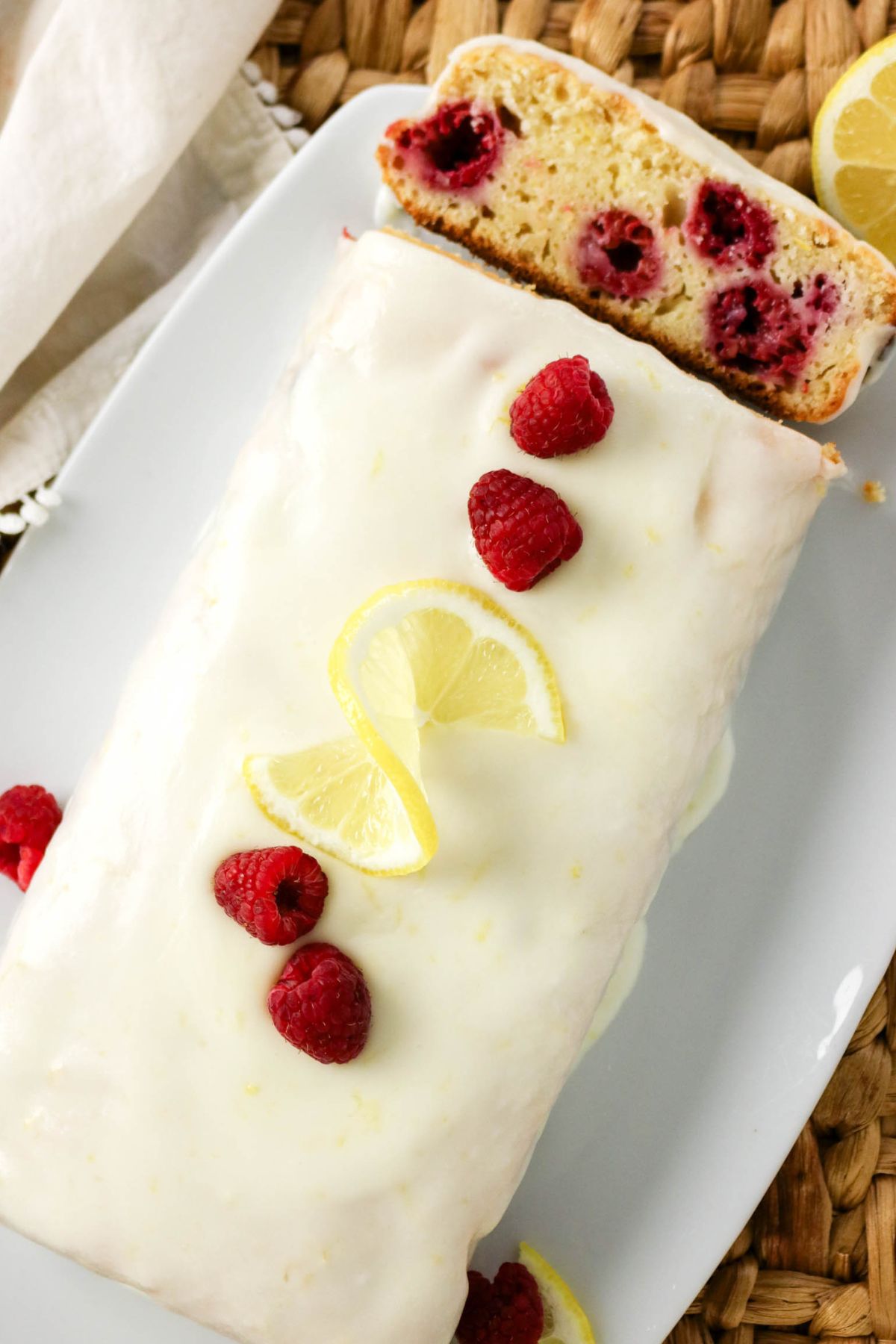 A lemon-glazed loaf cake topped with fresh raspberries and lemon slices, with a slice cut to show raspberries baked inside, on a white plate.