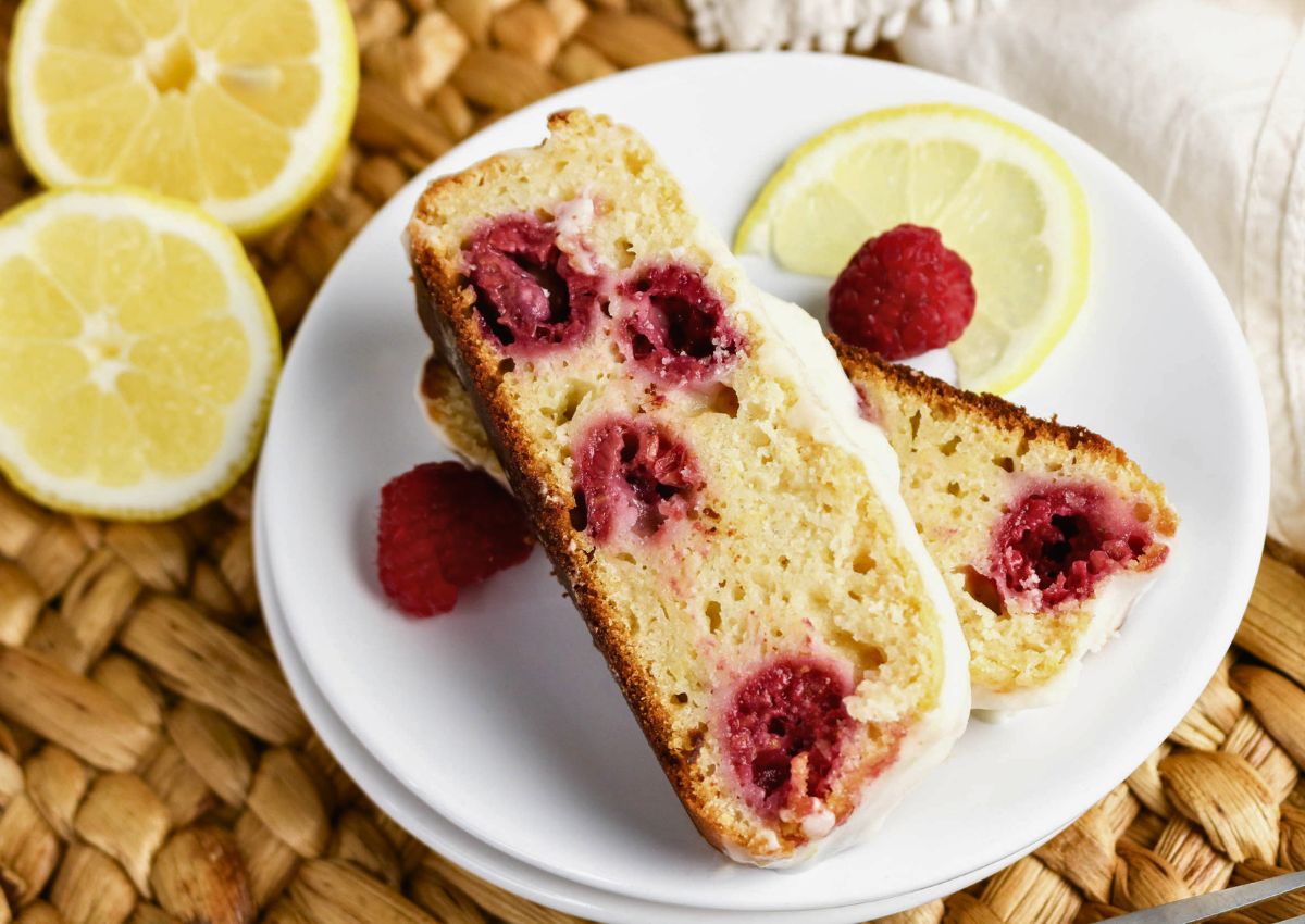 Two slices of raspberry lemon loaf cake on a white plate with fresh raspberries and lemon slices, displayed on a woven mat.