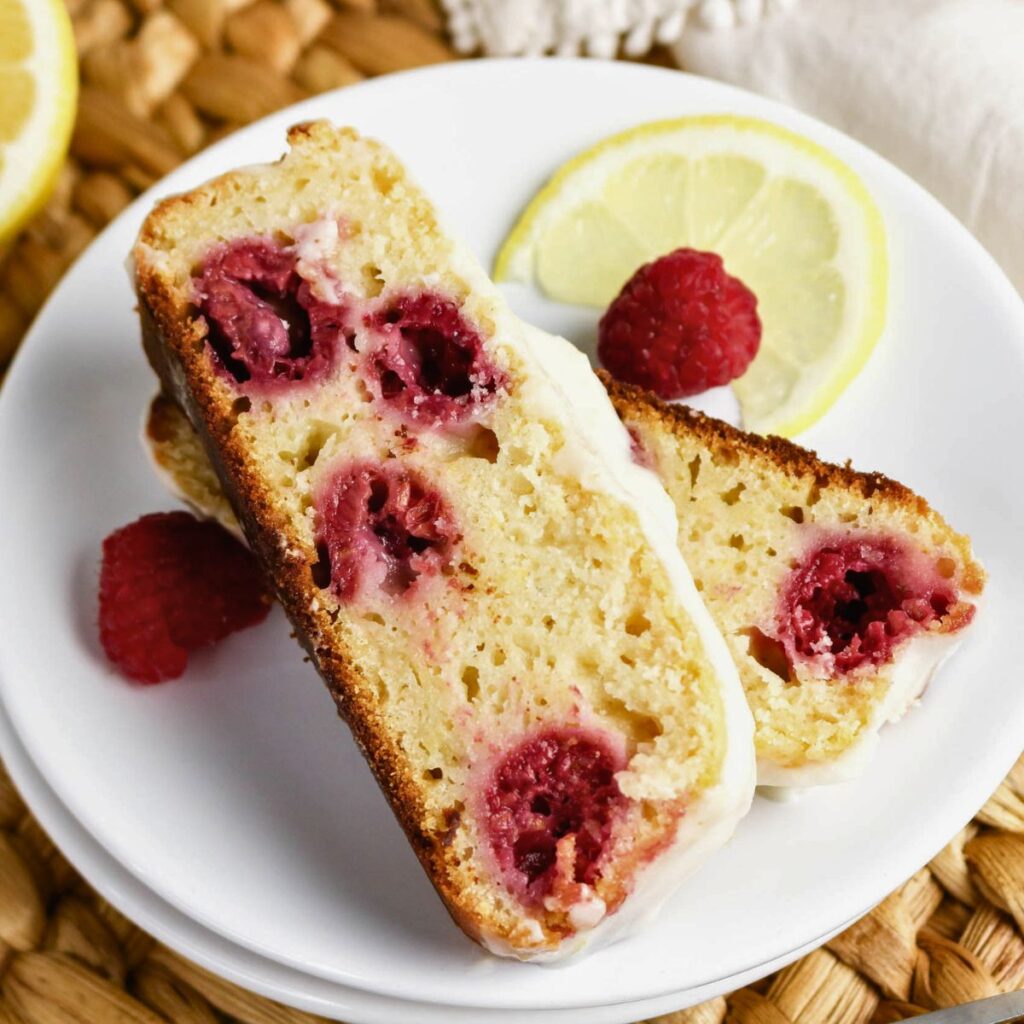 Two slices of raspberry lemon loaf cake with white icing on a white plate, garnished with fresh raspberries and a lemon slice.