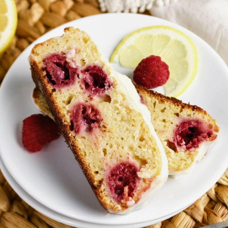 Two slices of raspberry lemon loaf cake with white icing on a white plate, garnished with fresh raspberries and a lemon slice.