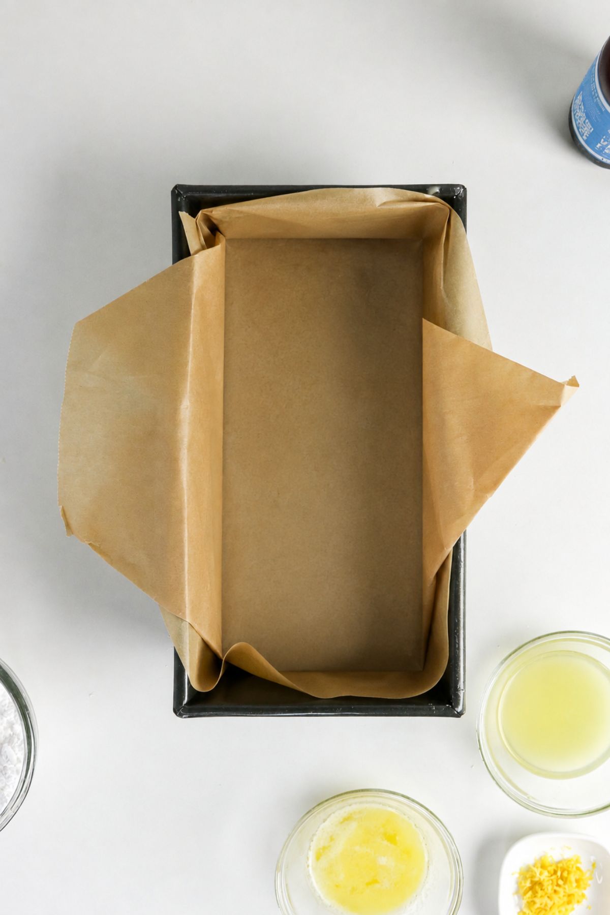 A rectangular loaf pan lined with parchment paper sits on a white surface, surrounded by small bowls of melted butter, lemon juice, lemon zest, and flour.
