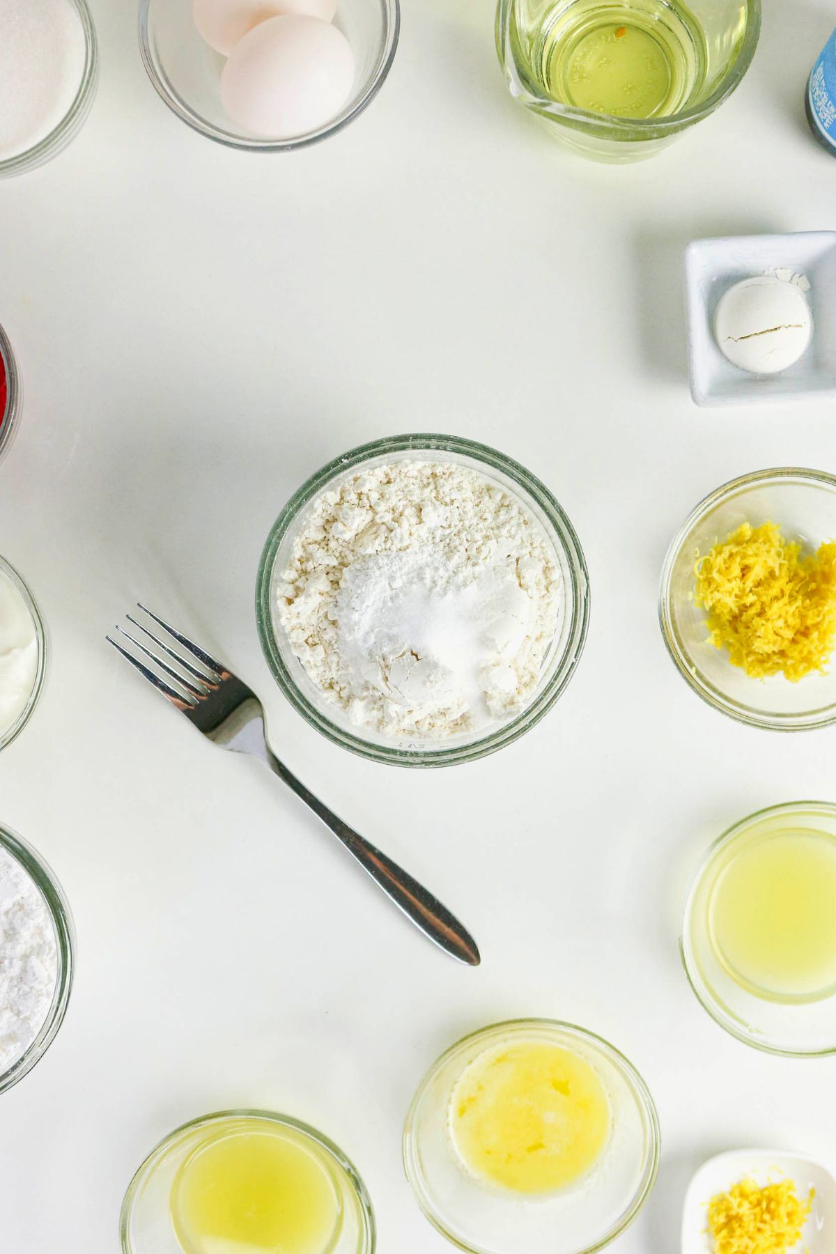 Various baking ingredients, including flour, eggs, lemon zest, lemon juice, and melted butter, are arranged in glass bowls on a white surface with a fork placed nearby.