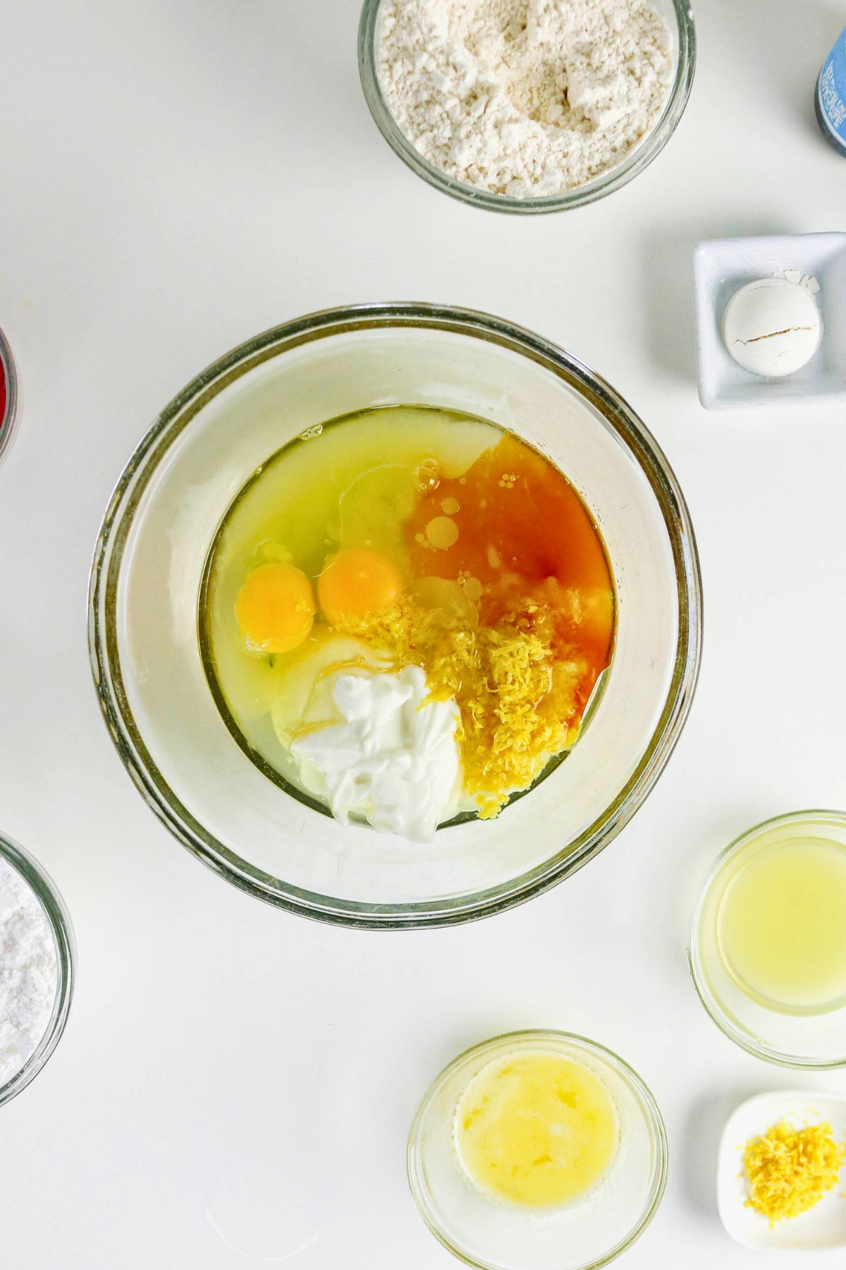 A mixing bowl with eggs, lemon zest, yogurt, and vanilla, surrounded by bowls of flour, sugar, lemon juice, butter, and a cracked egg shell.