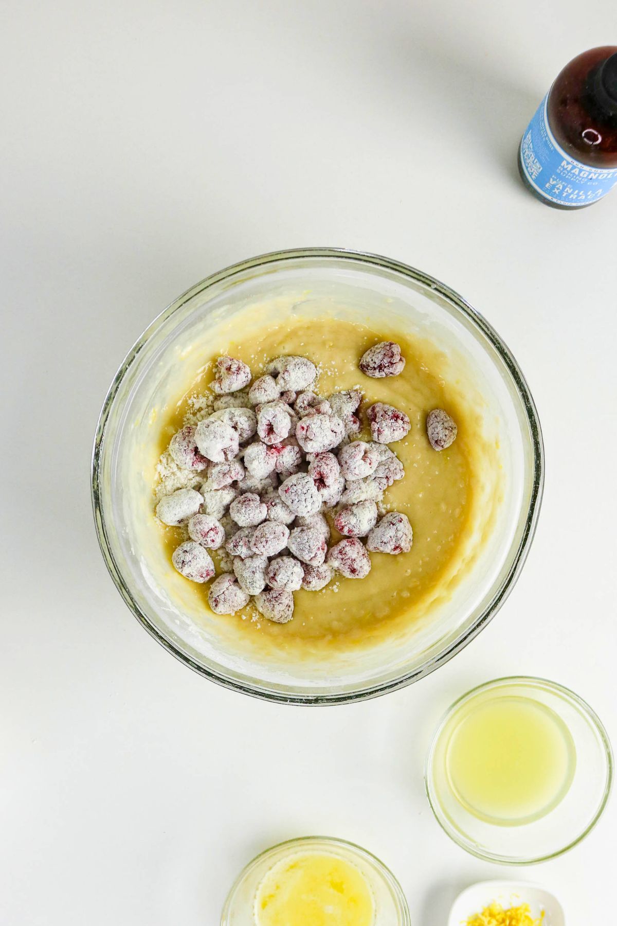 Glass bowl of batter with flour-dusted raspberries on top, surrounded by small bowls of melted butter, lemon juice, lemon zest, and a bottle of vanilla extract on a white surface.