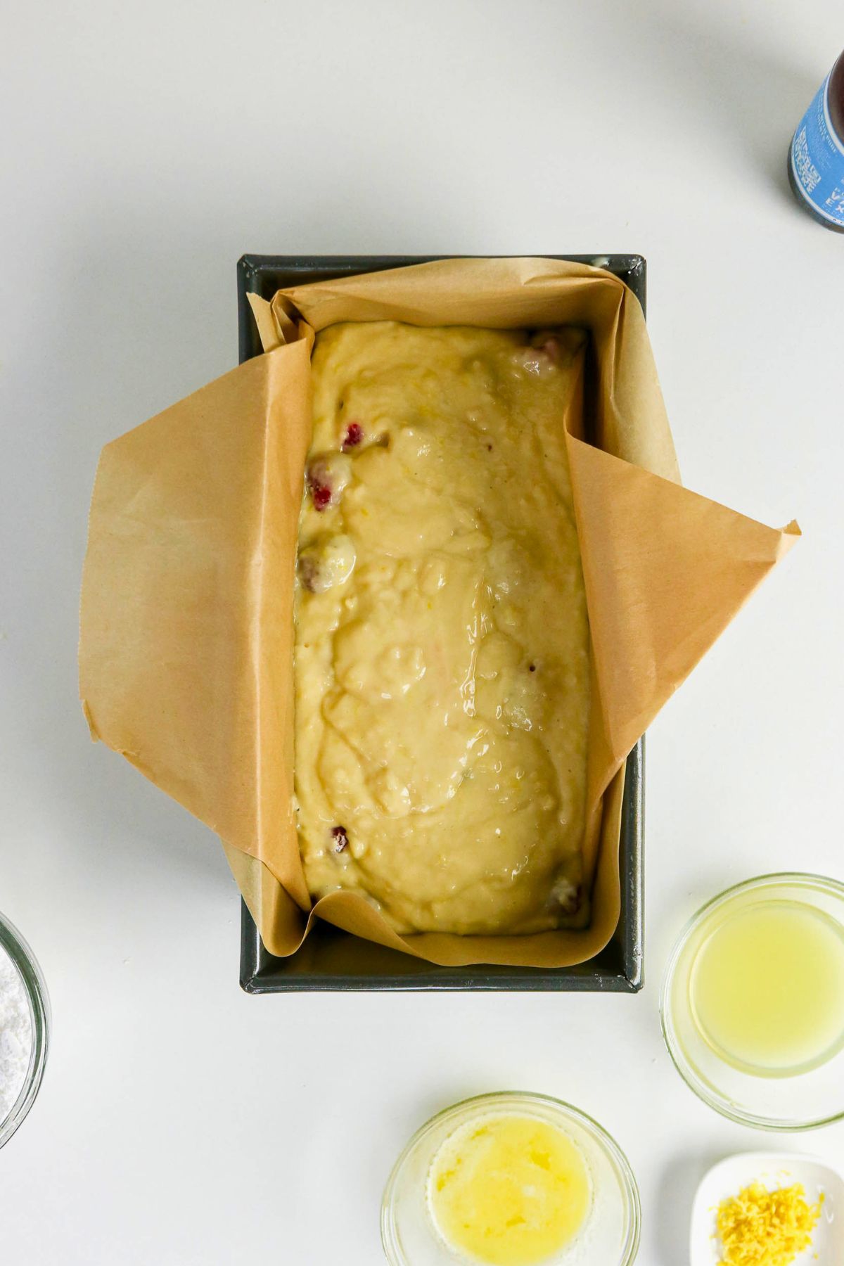 A loaf pan lined with parchment paper is filled with unbaked batter. Surrounding it are small bowls of melted butter, lemon juice, lemon zest, and a container of baking powder.