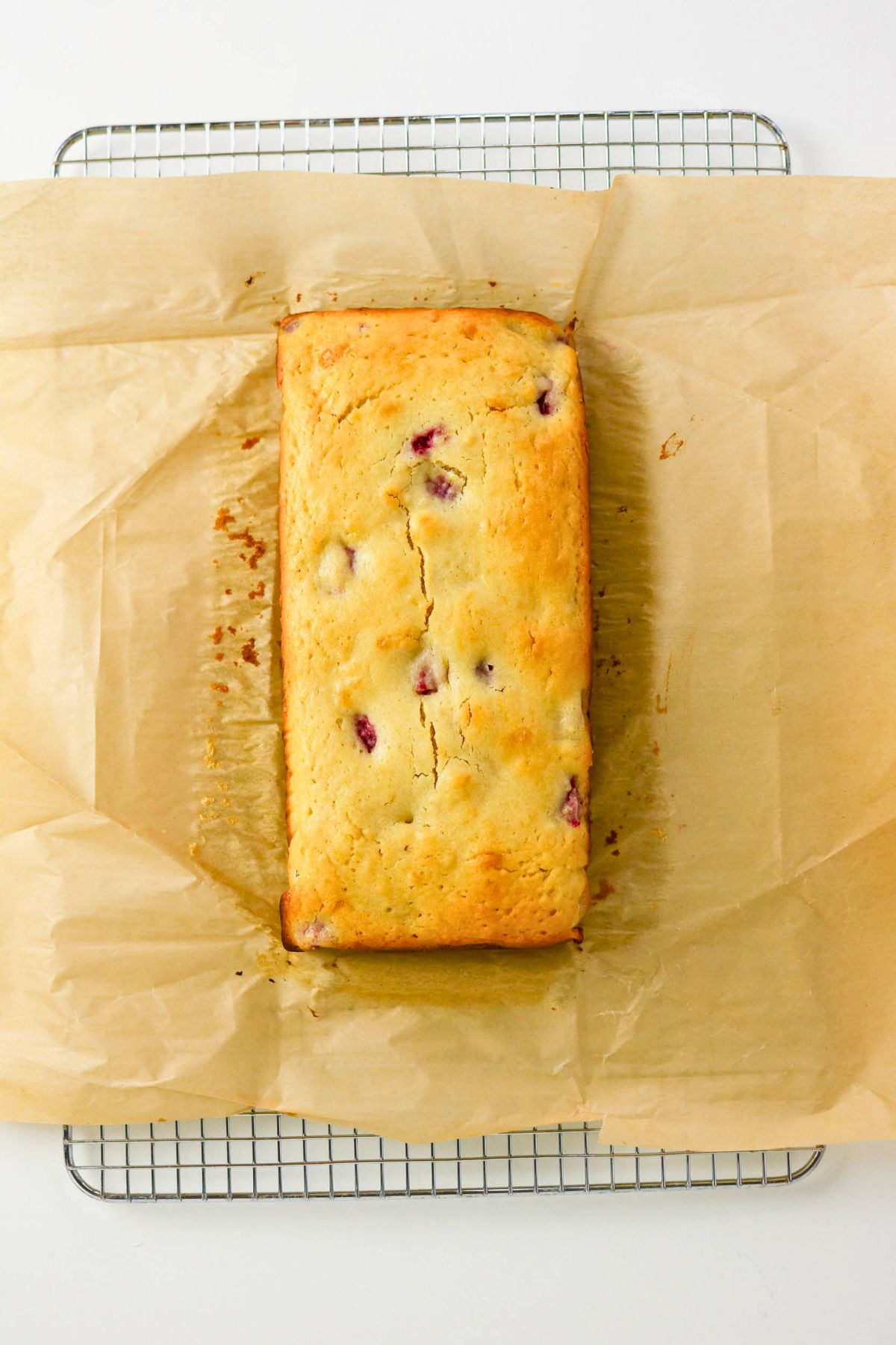 A golden brown loaf with visible berry pieces sits on parchment paper atop a cooling rack.