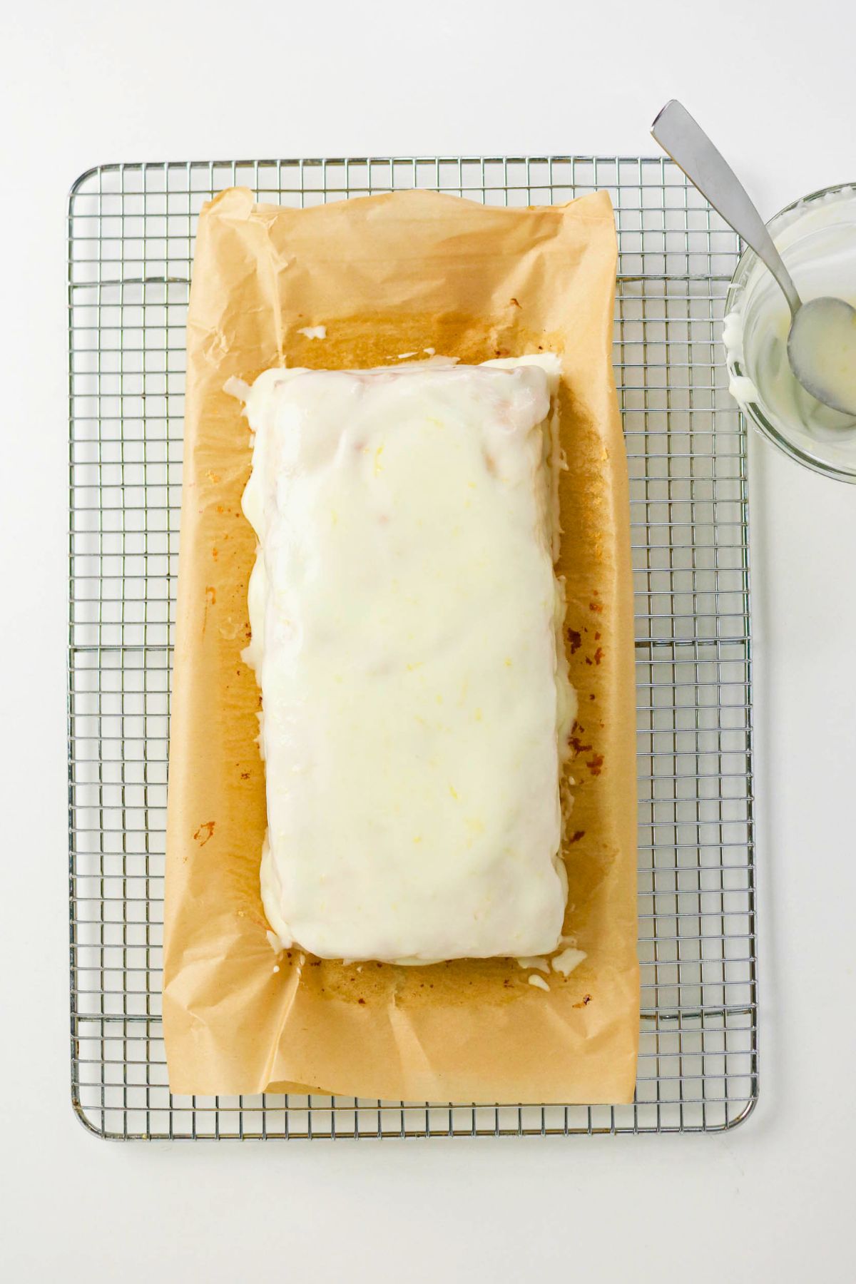 A rectangular loaf cake with white icing sits on parchment paper atop a cooling rack, with a spoon and glass bowl containing extra icing nearby.