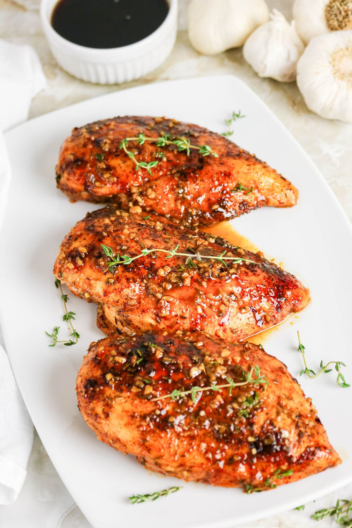 Three glazed chicken breasts garnished with herbs are arranged on a white rectangular plate, with a small bowl of dark sauce and heads of garlic in the background.
