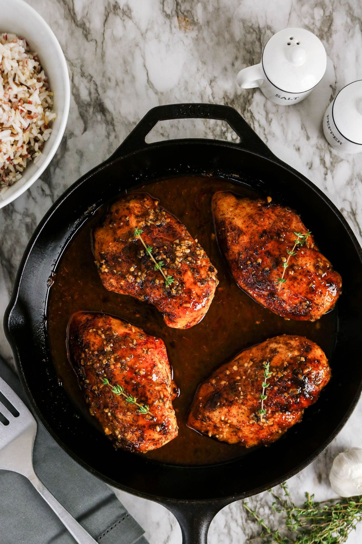 Four seasoned, cooked chicken breasts in a cast iron skillet with sauce, garnished with herbs. A bowl of rice and salt and pepper shakers are nearby on a marble surface.