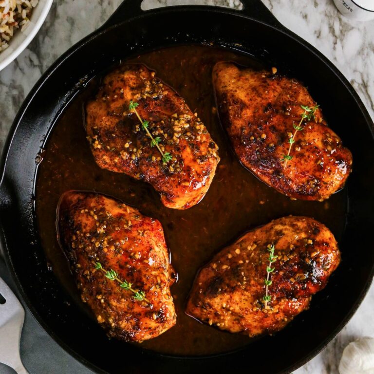 Four cooked, seasoned chicken breasts with garnish sit in a dark sauce inside a black cast iron skillet on a marble surface.