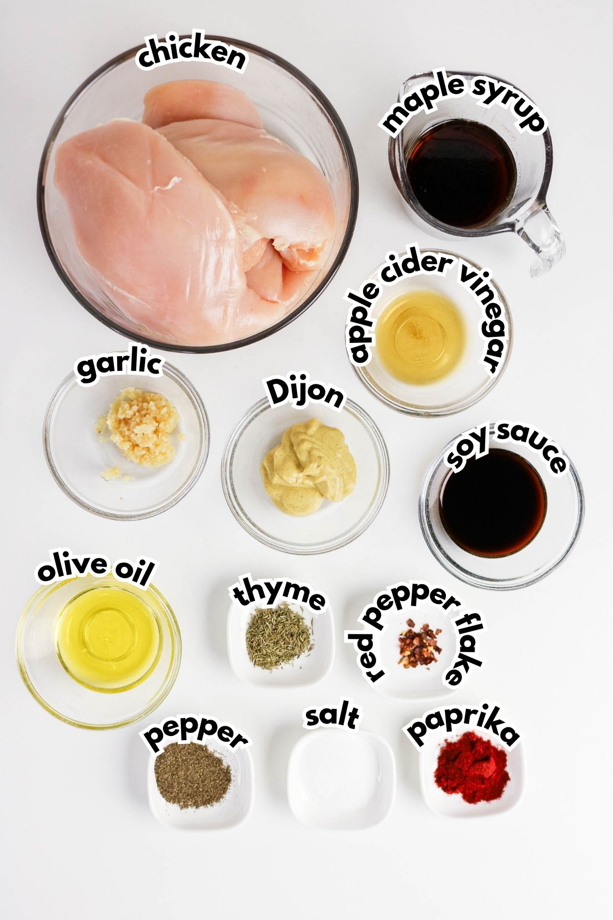 Raw chicken breast, maple syrup, apple cider vinegar, garlic, Dijon mustard, soy sauce, olive oil, thyme, red pepper flakes, paprika, pepper, and salt arranged in bowls on a white background.