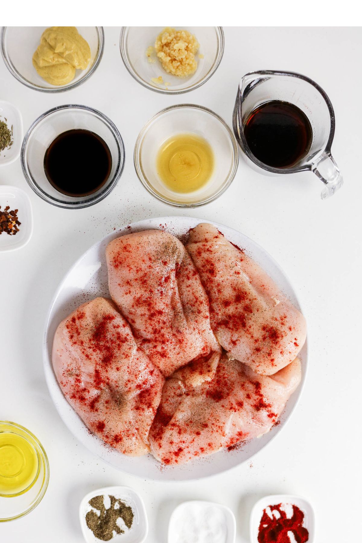 Raw chicken breasts seasoned with spices on a plate, surrounded by small bowls containing mustard, minced garlic, soy sauce, oil, vinegar, and various seasonings on a white background.