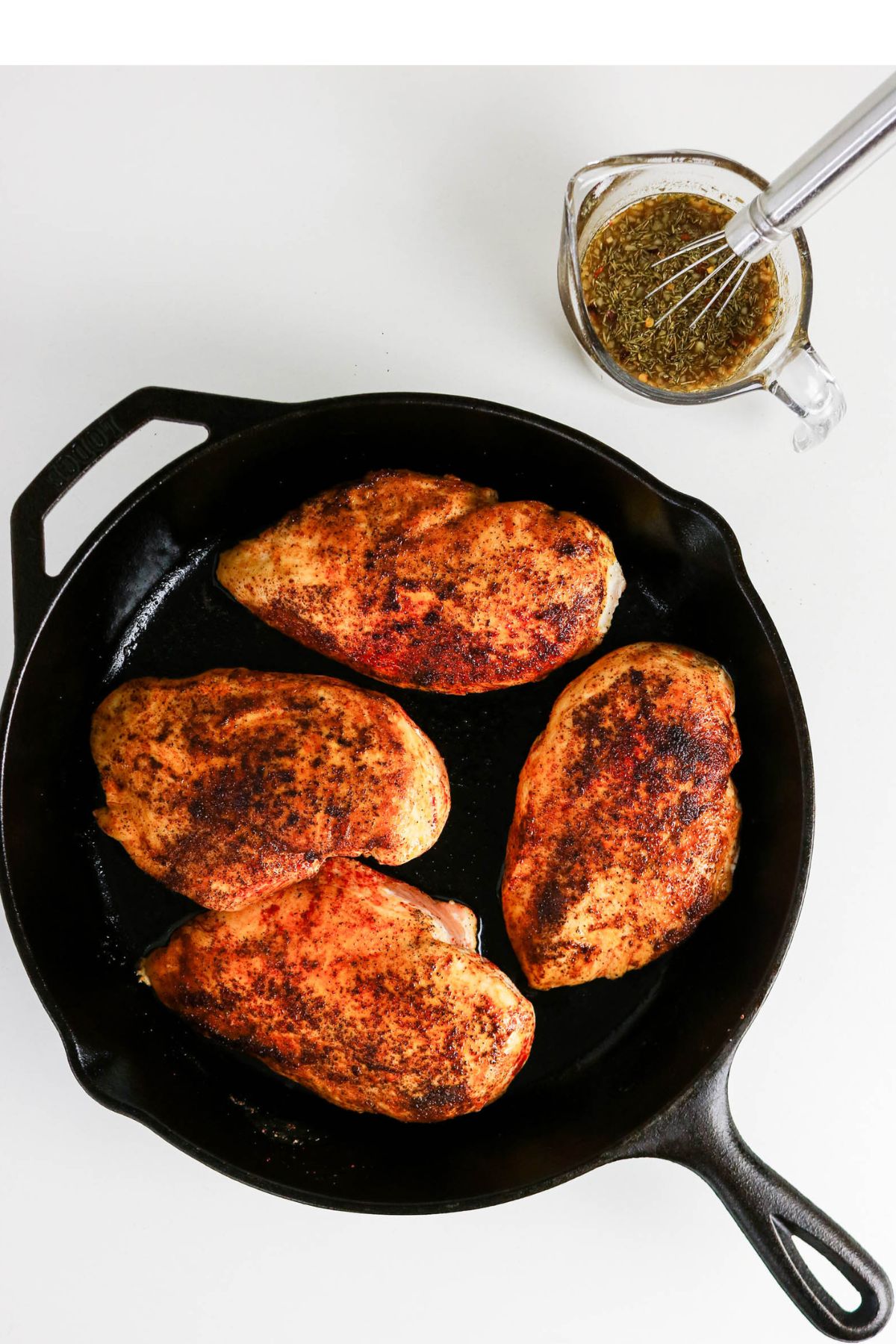 Four seasoned chicken breasts are cooking in a black cast iron skillet, with a small glass pitcher of herb marinade and a whisk placed nearby on a white surface.