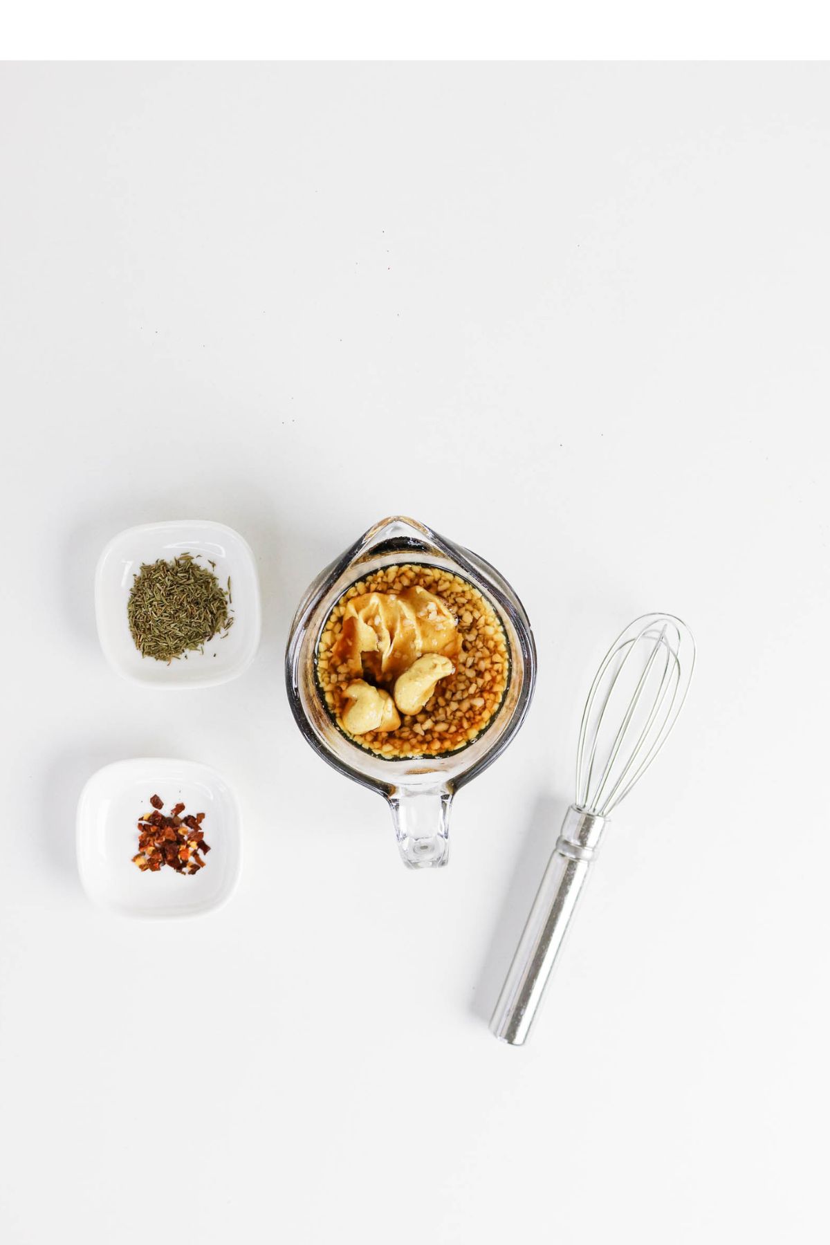 A glass measuring cup with mustard, garlic, and lentils, alongside two small dishes of herbs and red pepper flakes, and a metal whisk on a white background.