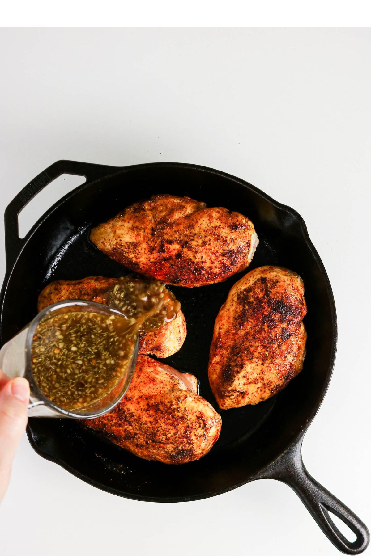 Four seasoned chicken breasts in a cast iron skillet, with herb sauce being poured over them from a glass container.