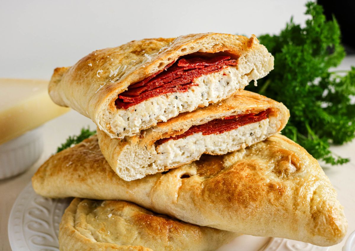 Two calzones are stacked on a white plate, each filled with ricotta cheese and slices of pepperoni. Fresh parsley is visible in the background.