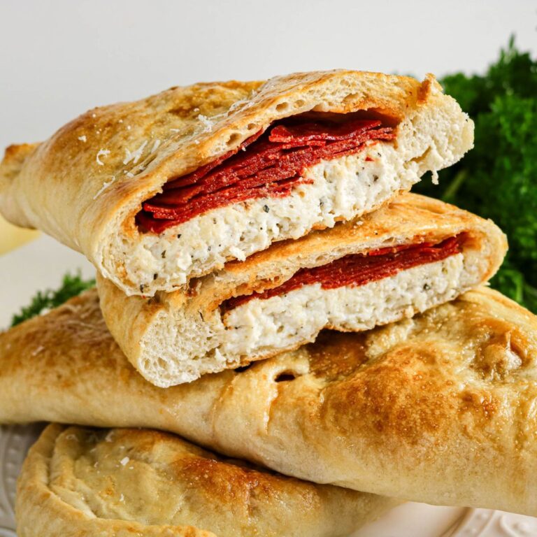A calzone cut in half, showing layers of melted cheese and pepperoni inside baked golden-brown dough, with leafy greens in the background.