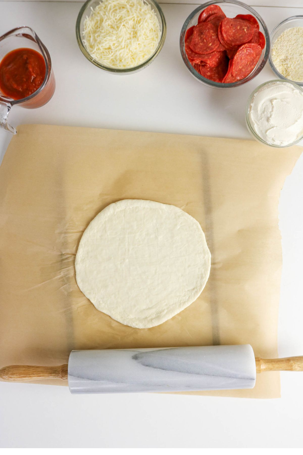 Pizza dough rolled out on parchment paper with a marble rolling pin, surrounded by bowls of sauce, shredded cheese, pepperoni, and other pizza toppings.