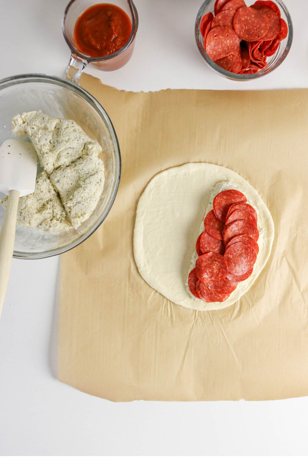 Pizza dough on parchment paper topped with rows of pepperoni; nearby bowls hold pizza sauce, sliced pepperoni, and seasoned ricotta cheese.