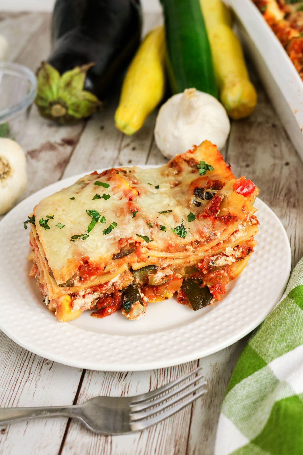A slice of vegetable lasagna topped with herbs sits on a white plate, with a fork beside it and fresh vegetables in the background.