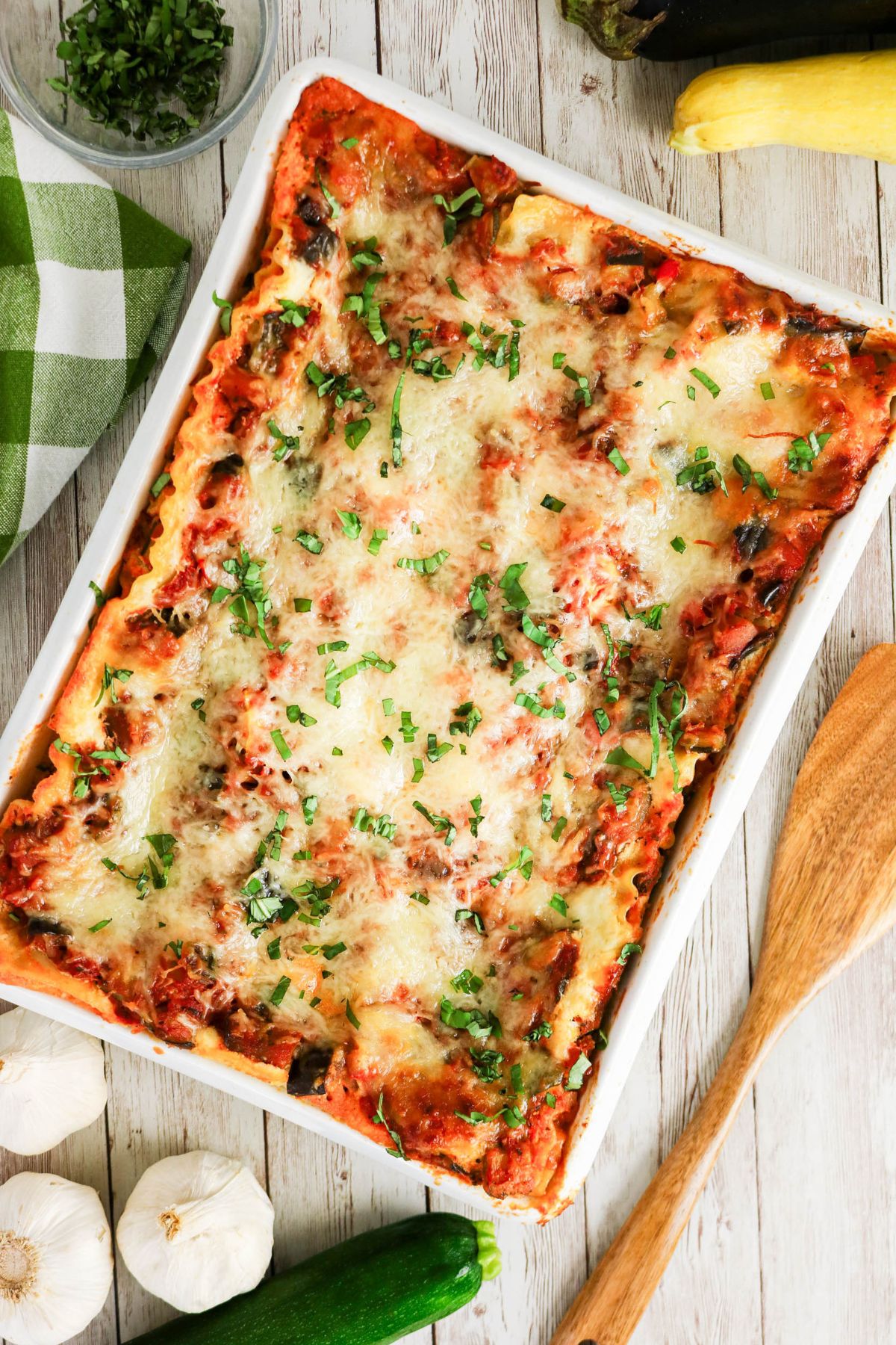 Baked lasagna in a rectangular dish topped with melted cheese and herbs, surrounded by fresh vegetables, garlic, a wooden spoon, and a green-checked cloth.