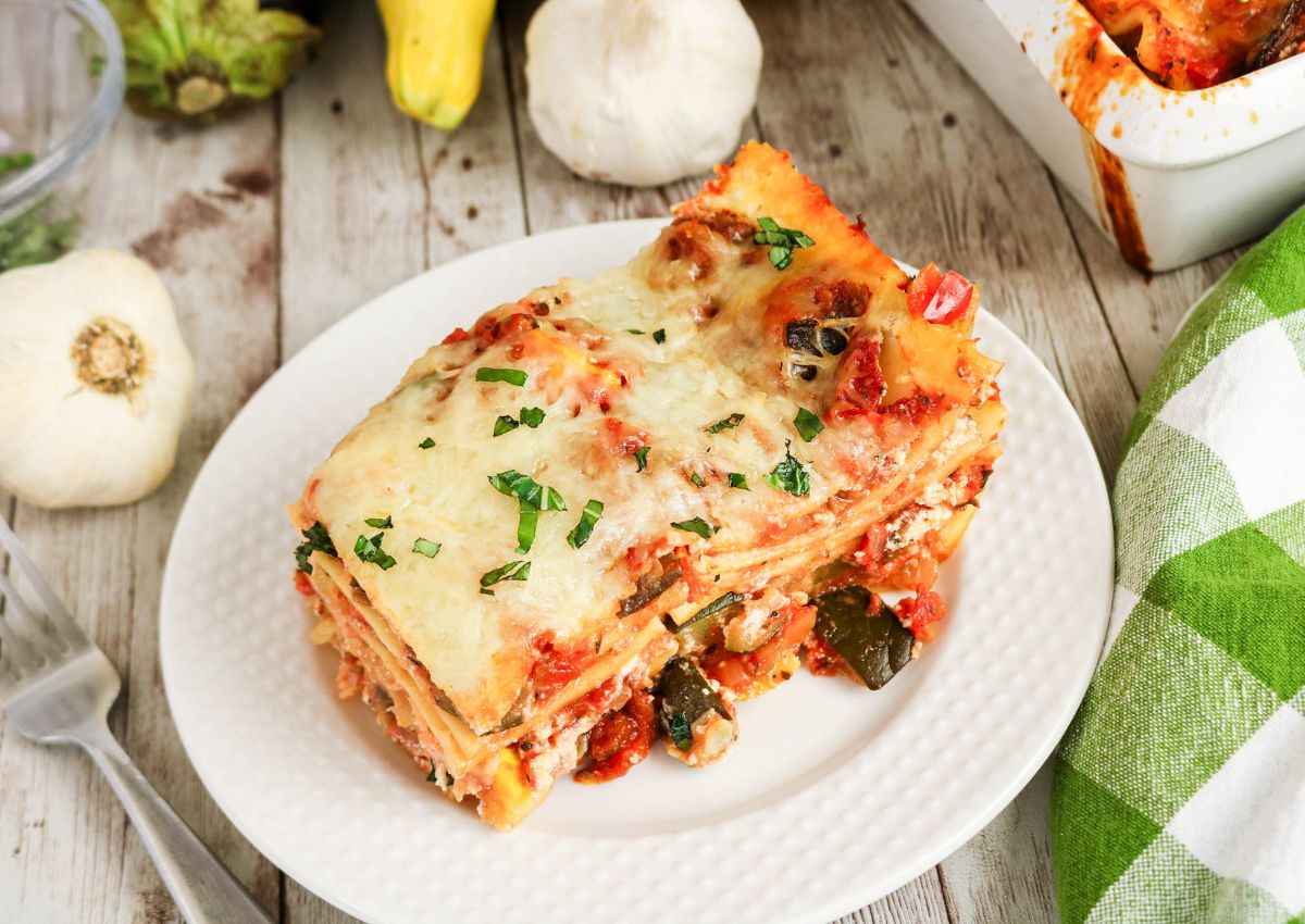 A slice of vegetable lasagna topped with melted cheese and herbs is served on a white plate with garlic and vegetables in the background.