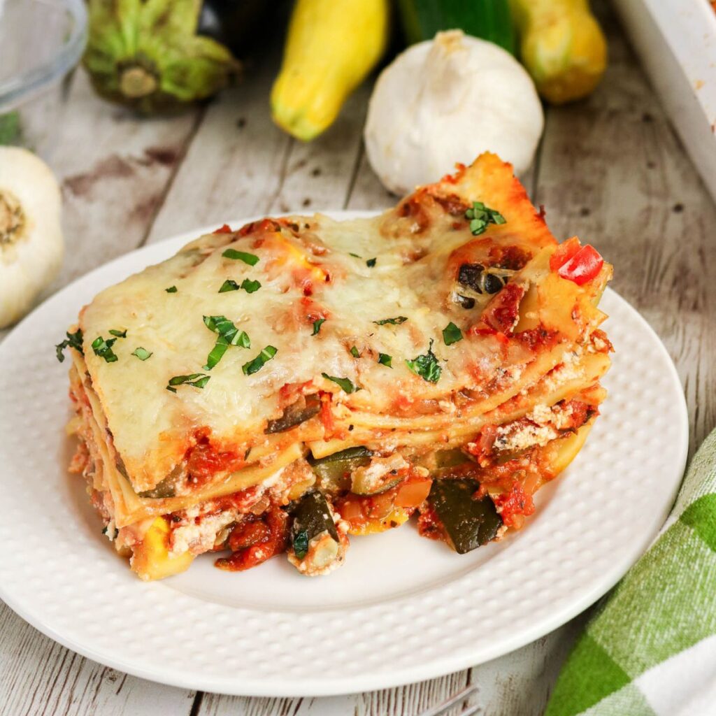 A slice of vegetable lasagna with melted cheese and herbs on a white plate, surrounded by fresh garlic and squash on a wooden table.