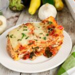 A slice of vegetable lasagna with melted cheese and herbs on a white plate, surrounded by fresh garlic and squash on a wooden table.