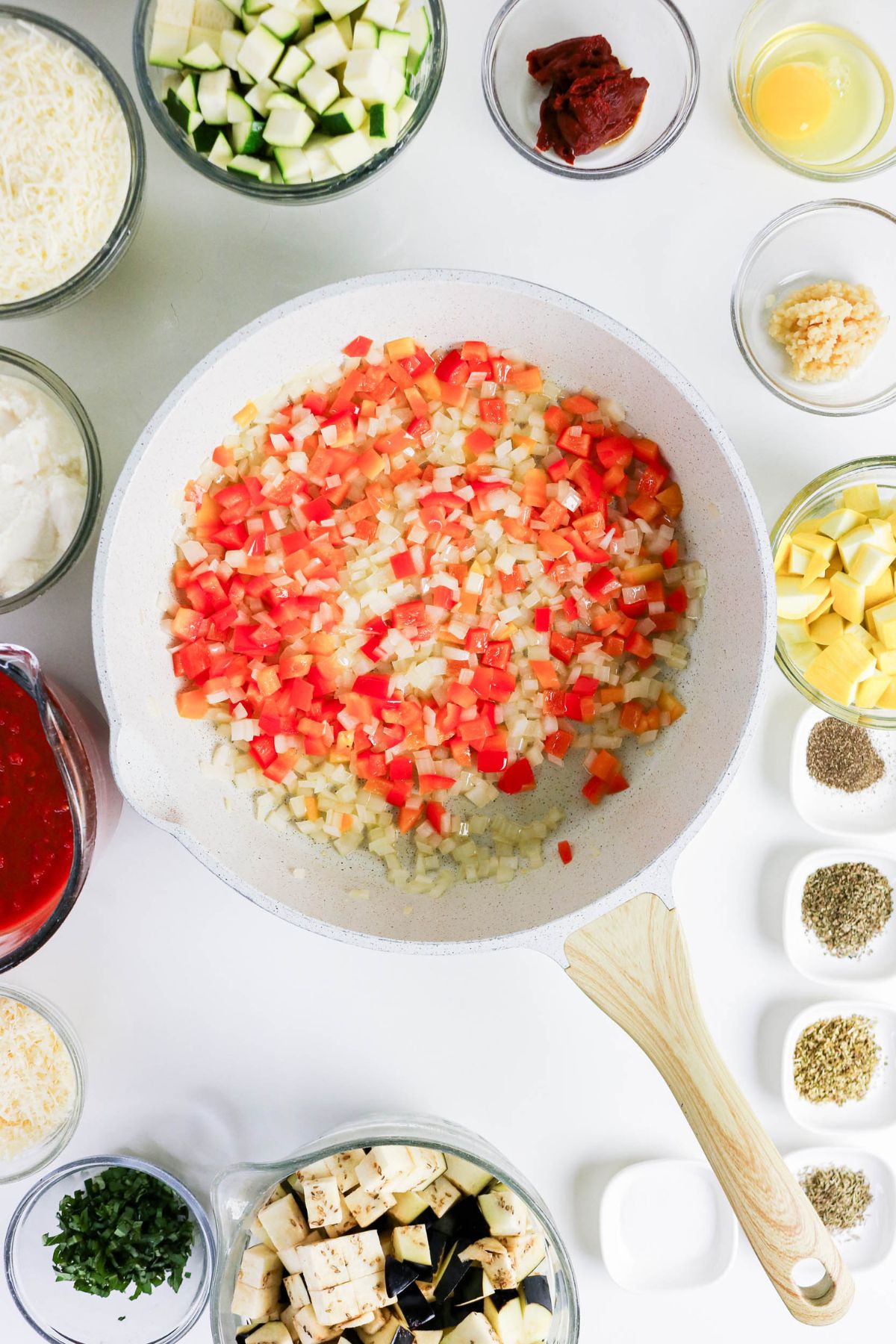 Chopped onions and red bell peppers cooking in a white skillet, surrounded by bowls of diced vegetables, spices, herbs, cheese, sauce, and other ingredients on a white surface.