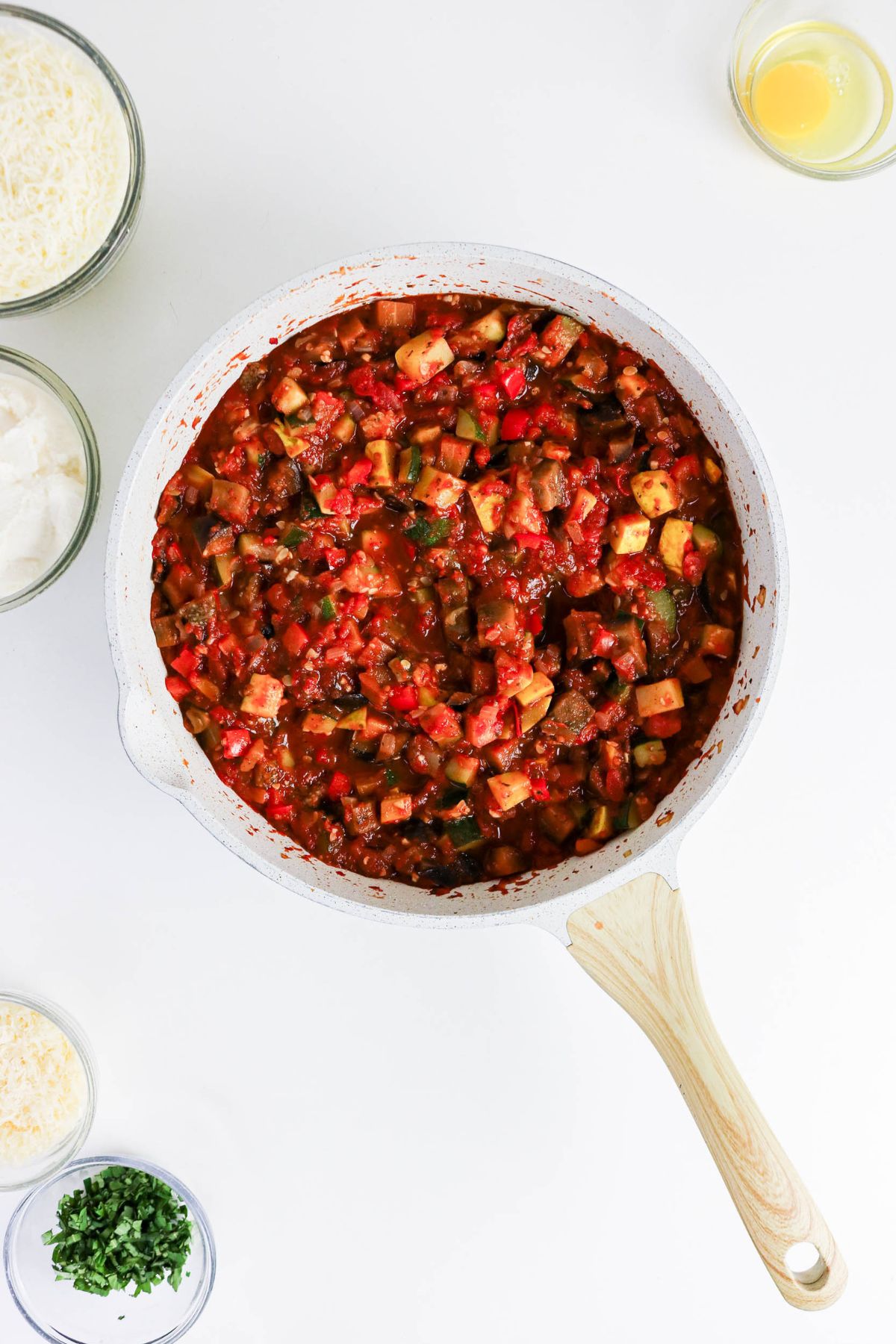 A white skillet filled with chopped vegetables in tomato sauce is on a white surface, surrounded by bowls of cheese, herbs, and a cracked egg.