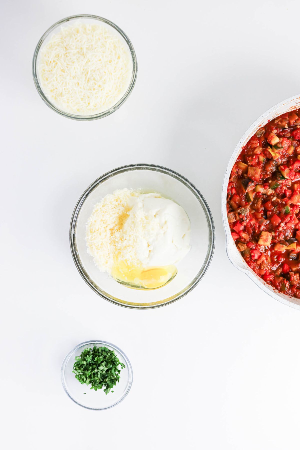 Three bowls with shredded cheese, ricotta mixture with egg, and chopped herbs next to a pan of tomato-based vegetable sauce on a white surface.